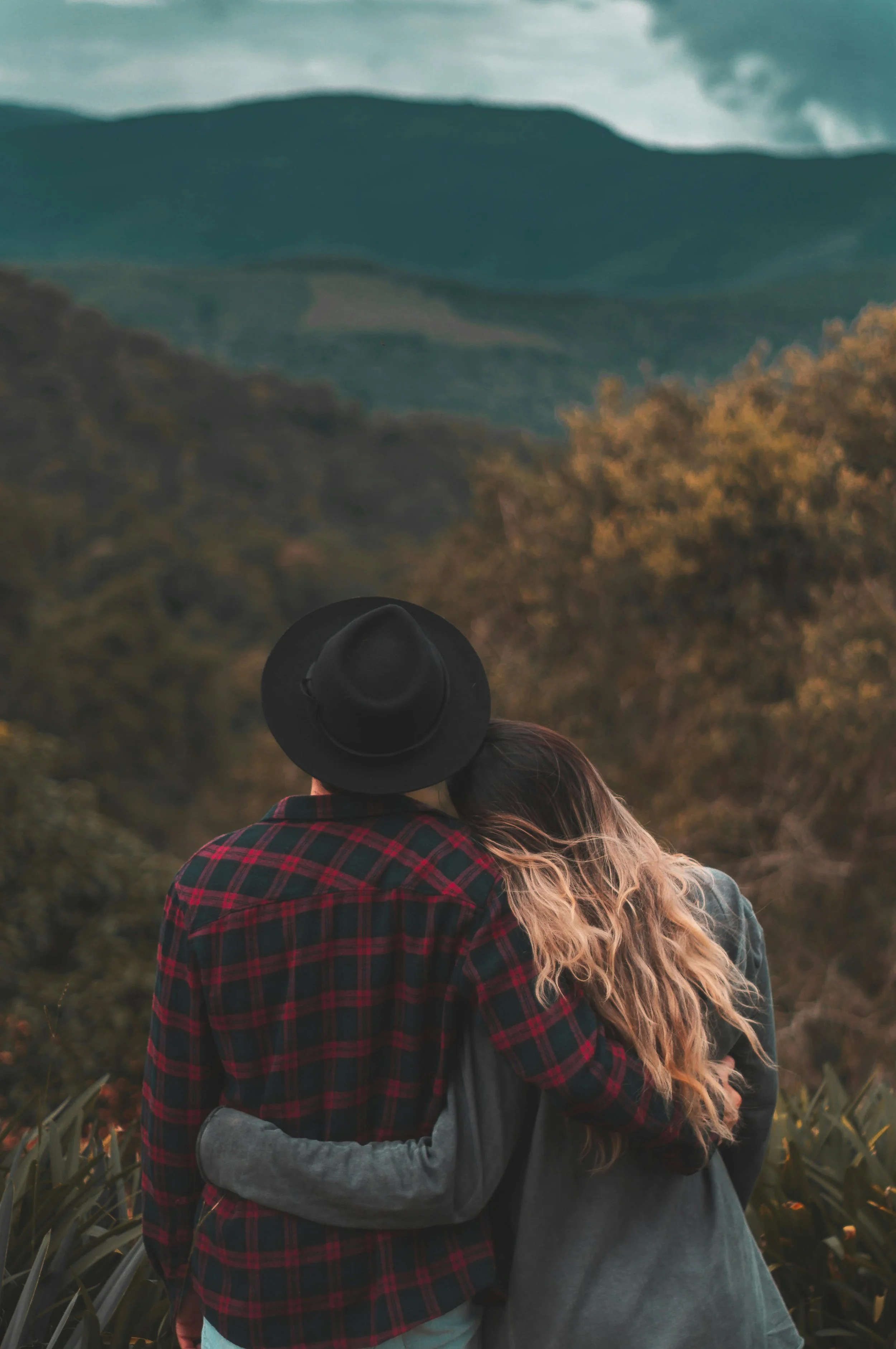 Una pareja abrazada en un paisaje natural con montañas y árboles en el fondo.
