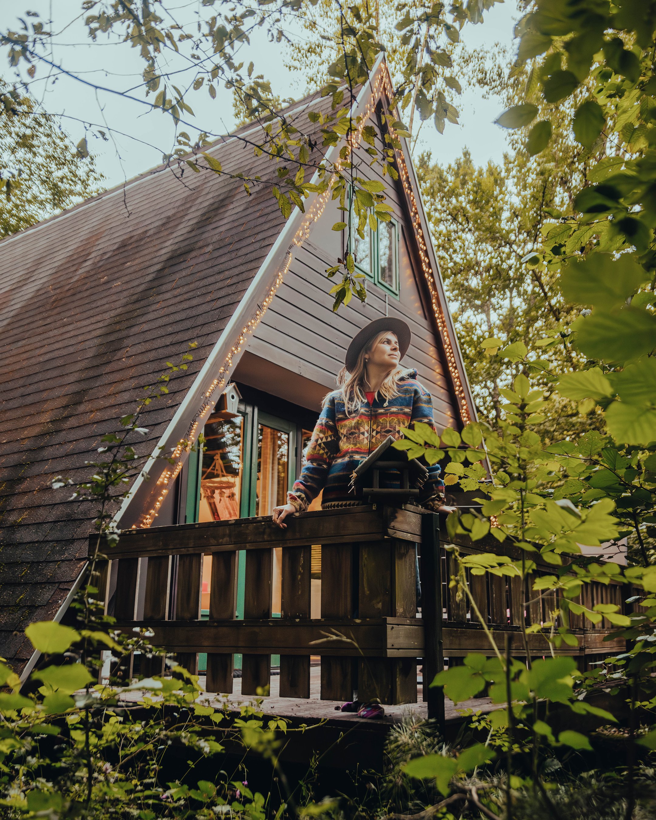 A woman standing on the balcony of an A-frame cabin decorated with string lights, surrounded by trees and greenery, looking thoughtfully into the distance.