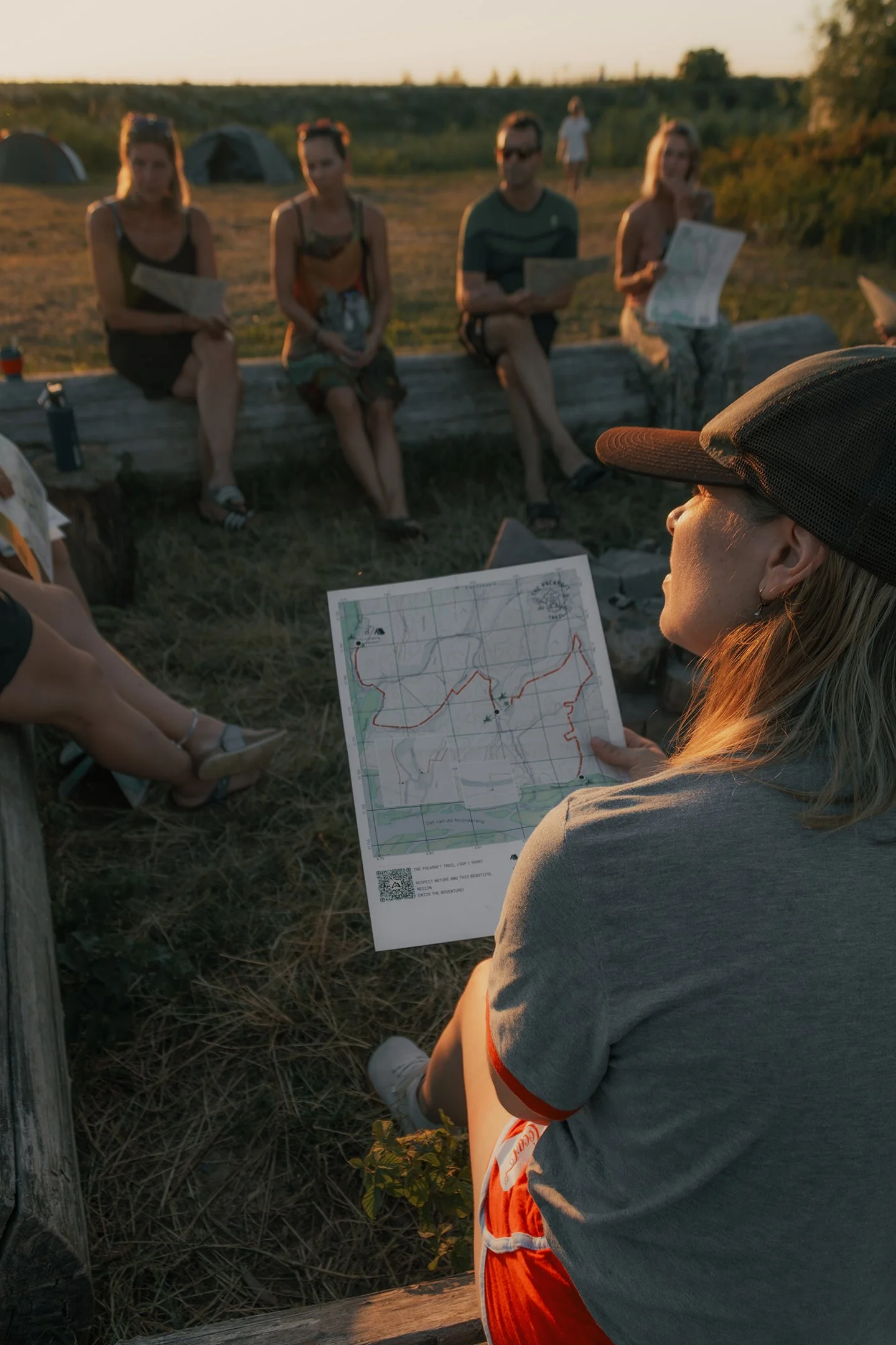 A group of people sitting outdoors in a circle, with a woman in the foreground holding a map, likely during a camping or outdoor activity at sunset.