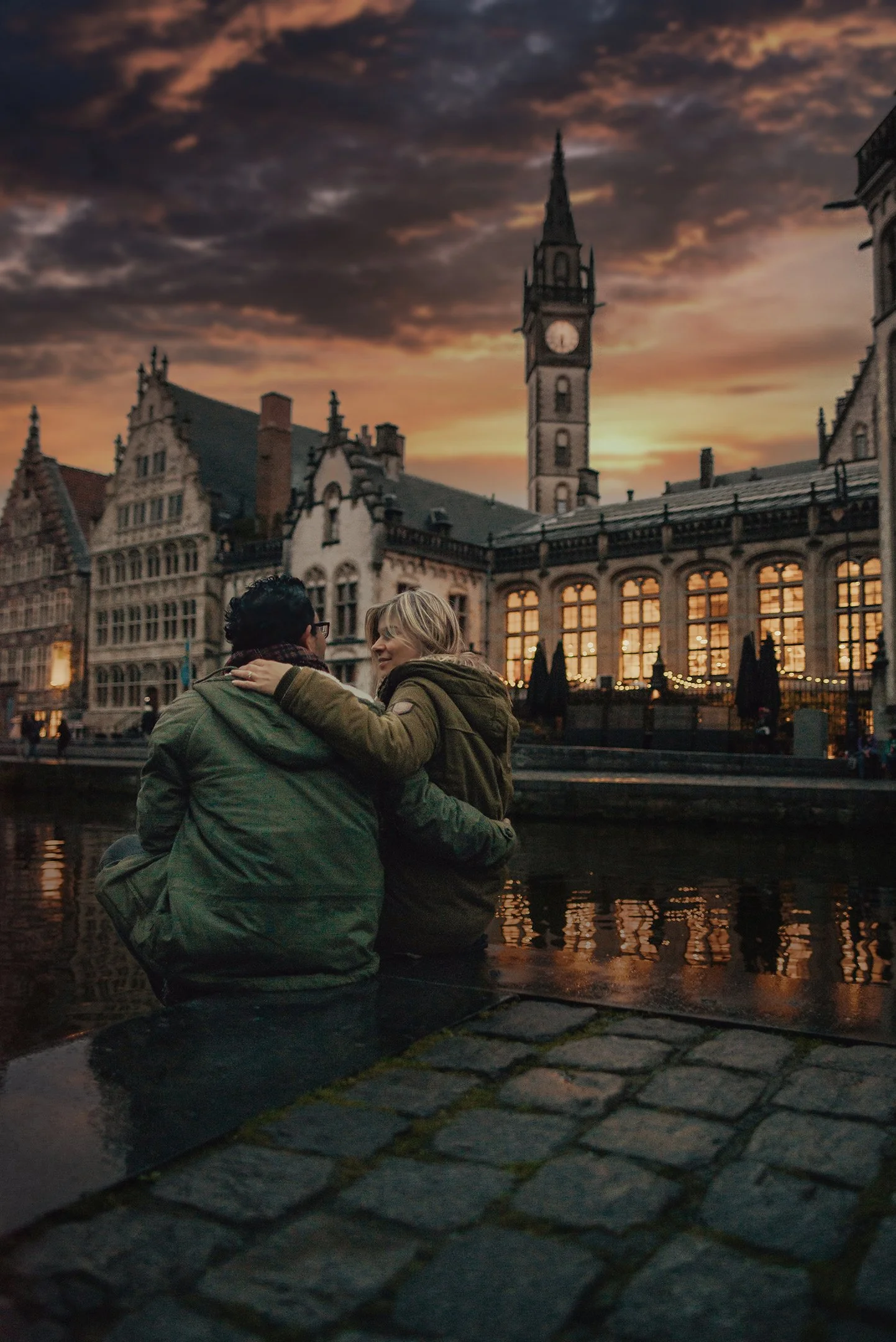 Una pareja sentada junto a un canal en una ciudad europea durante el atardecer, con edificios históricos y un reloj en la torre al fondo.
