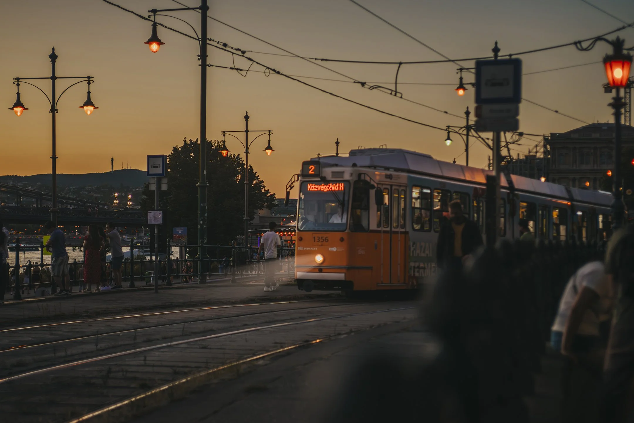 A tram arriving at a station during sunset, with hanging street lamps and people waiting on the platform.