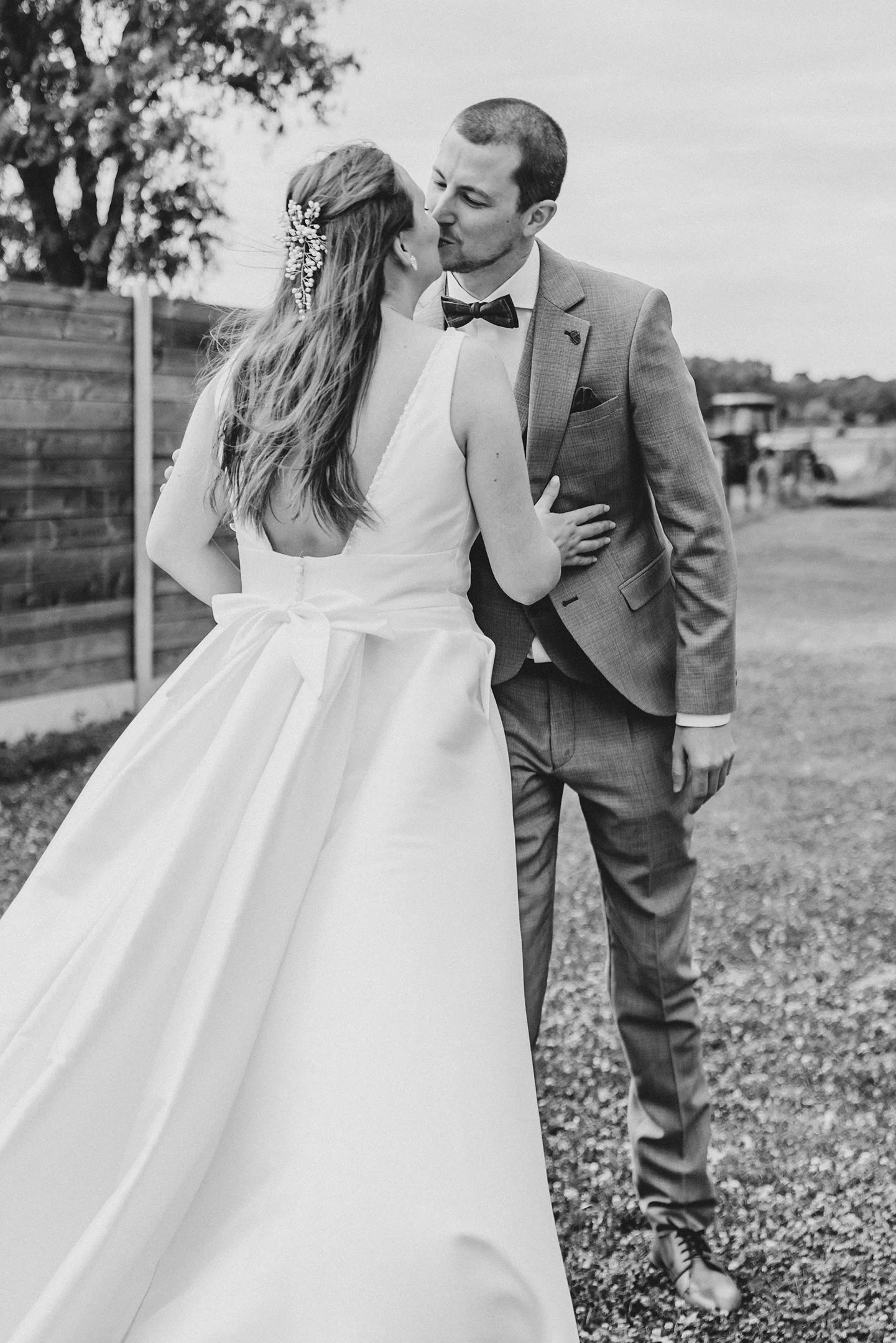 Una pareja de novios se besa en un entorno al aire libre durante su boda, ella usa un vestido largo y él lleva un traje con moño.