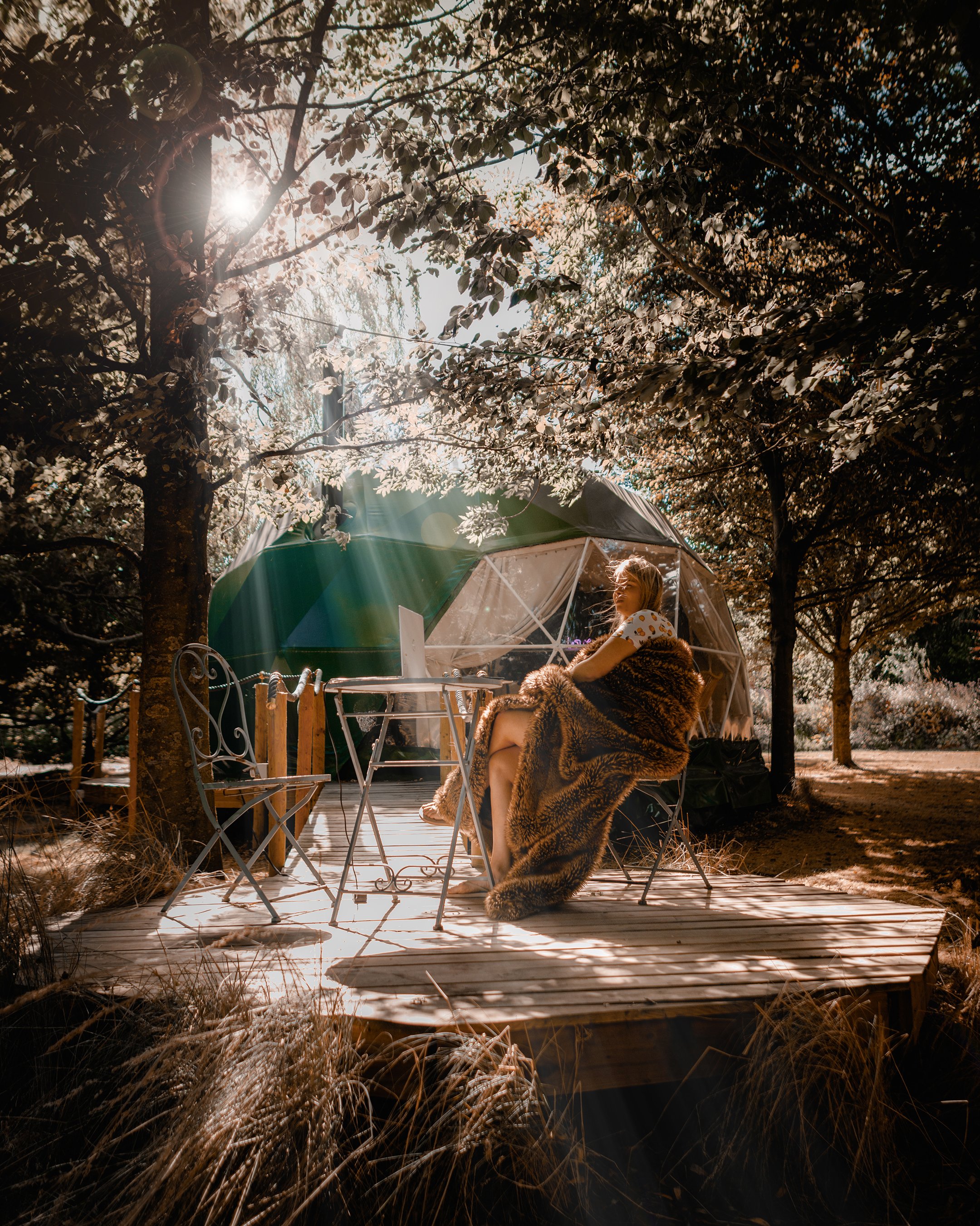 A woman sitting on a chair on a wooden deck, covered with a fur blanket, surrounded by trees with sunlight filtering through the leaves, and a geodesic dome tent in the background.