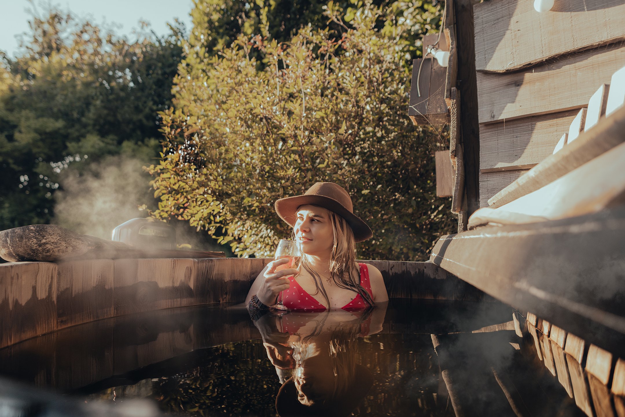 A woman in a red swimsuit and brown wide-brimmed hat relaxing in a wooden hot tub outdoors, holding a glass of wine, surrounded by greenery and sunlight.