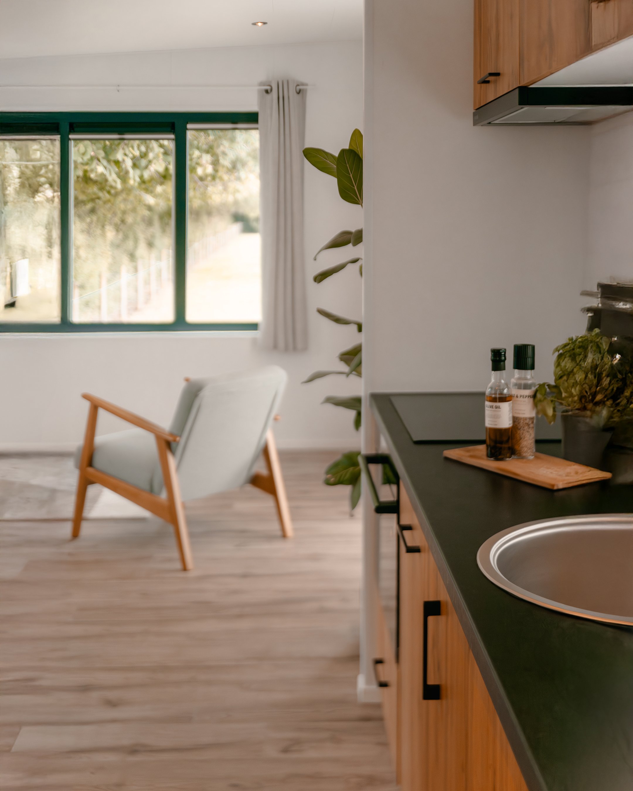 View of a cozy living room with large window, white curtains, wooden flooring, a light-colored armchair with wooden legs, and part of a kitchen with black countertop, wooden cabinets, and some bottles and plants on the counter.