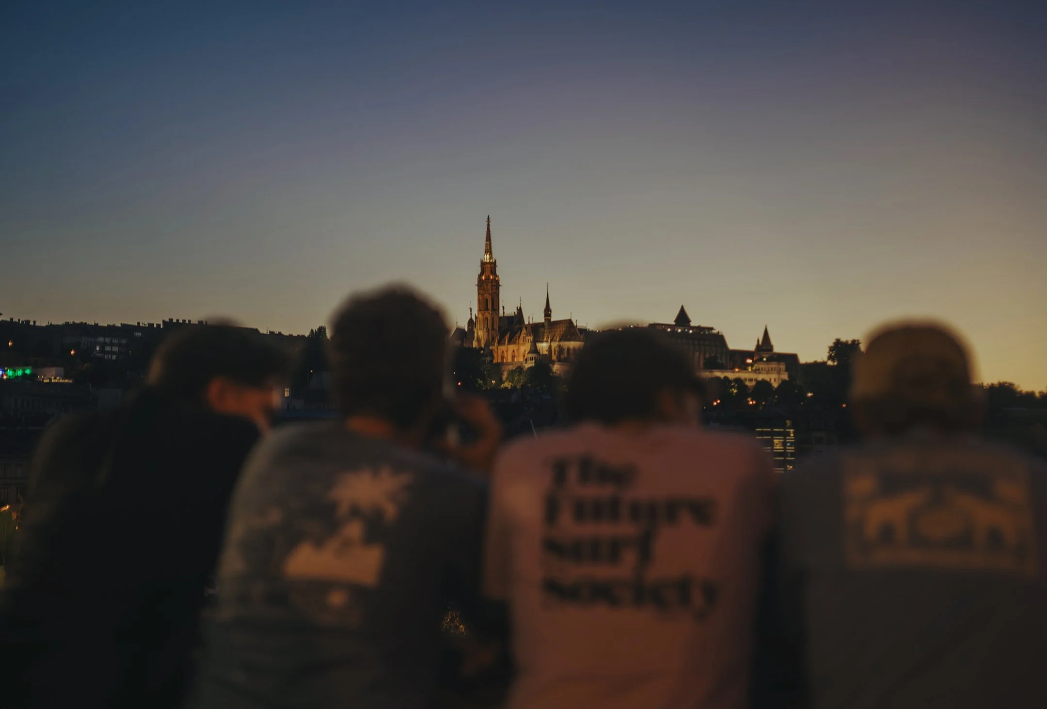 Four people sitting outdoors at sunset, overlooking a cityscape with a prominent church steeple in the distance.