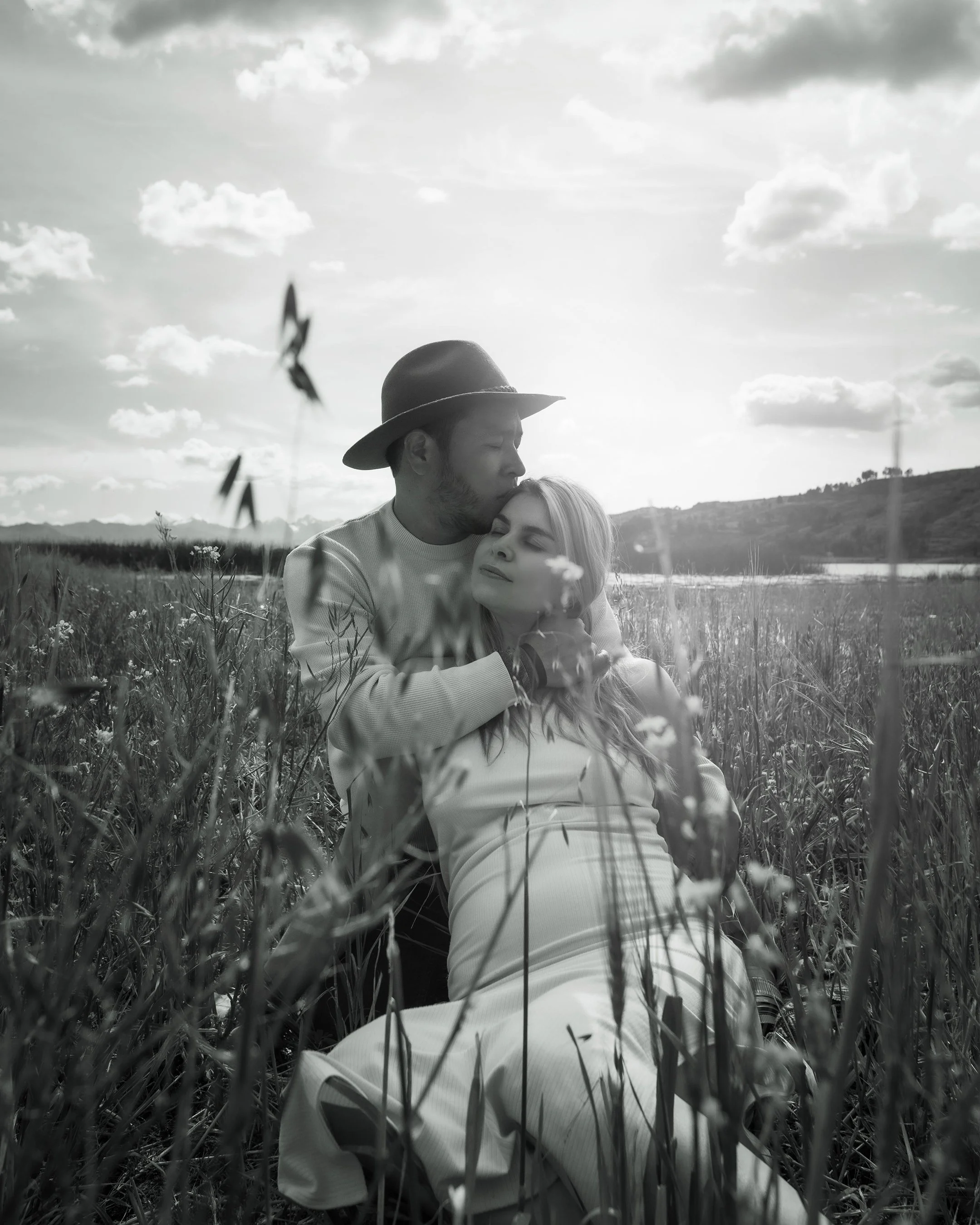 Pareja en un campo, él besando y abrazando a ella, en un ambiente natural con cielo nublado.