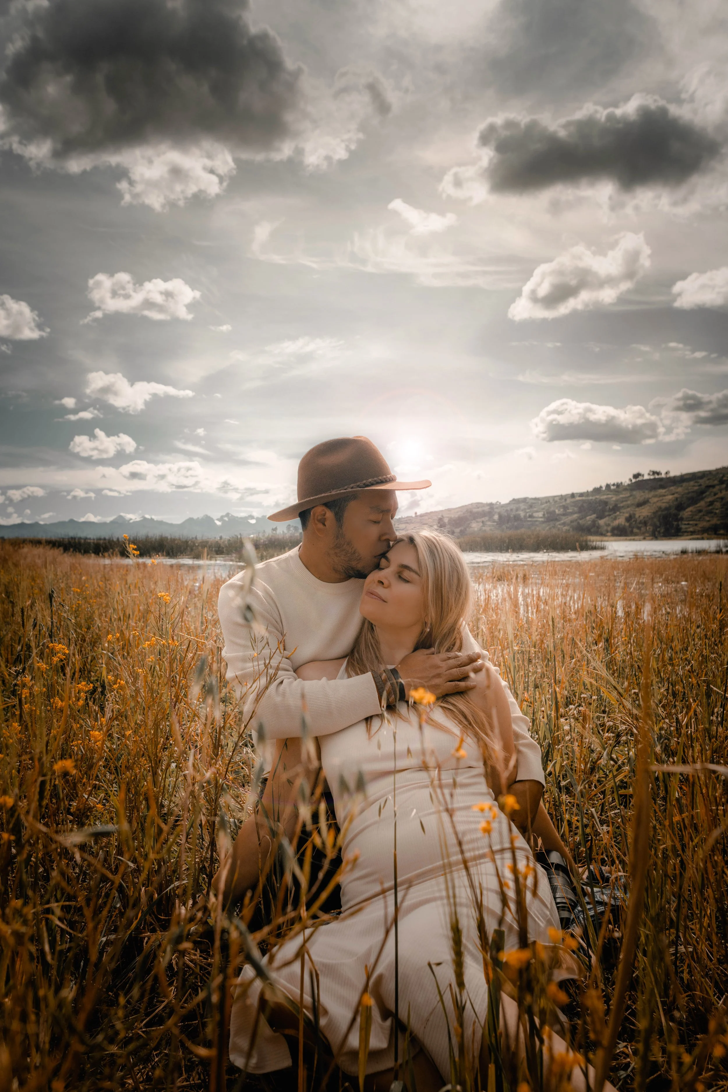 una pareja en un campo de flores en un día soleado, él beso en la frente a ella mientras ella está en el suelo, durante el atardecer con cielo nublado y montañas a la distancia