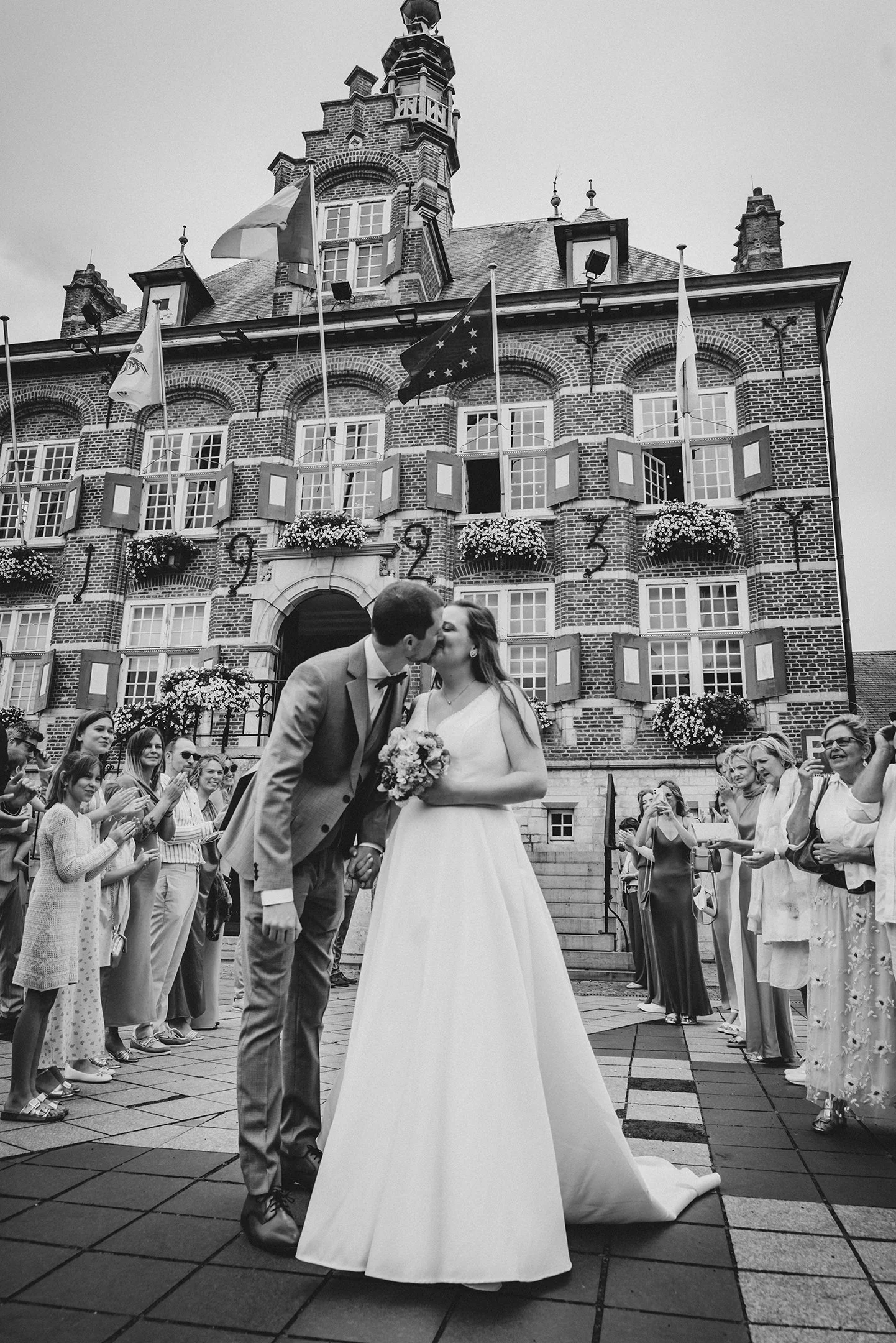 Una pareja de recién casados se besa en la frente en medio de una ceremonia al aire libre frente a un edificio de ladrillos con banderas y ventanas con plantas. La novia lleva vestido largo y el novio traje gris. Hay varias personas a su alrededor to