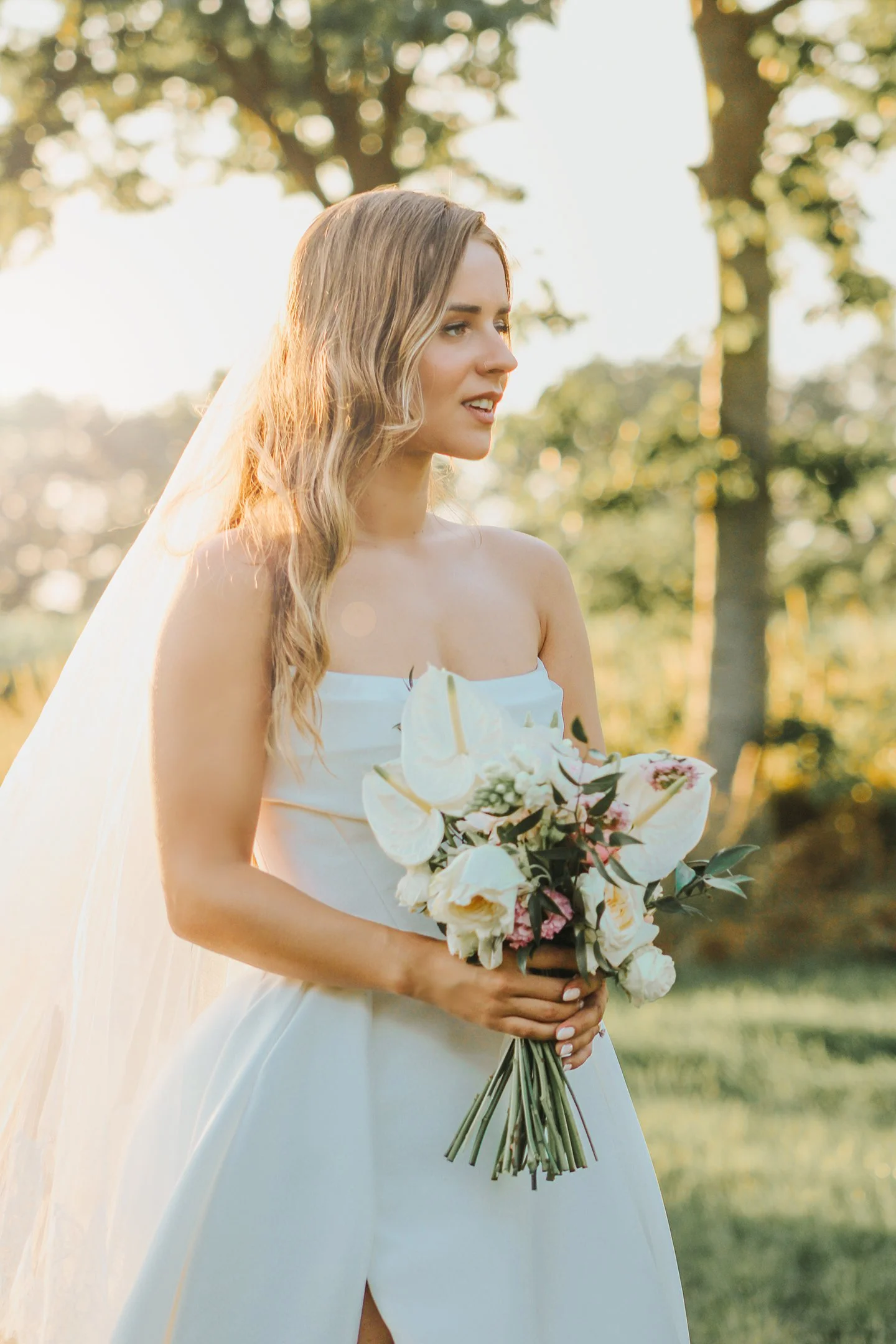 Mujer con vestido de novia sosteniendo ramo de flores blancas en un entorno de naturaleza durante el atardecer.