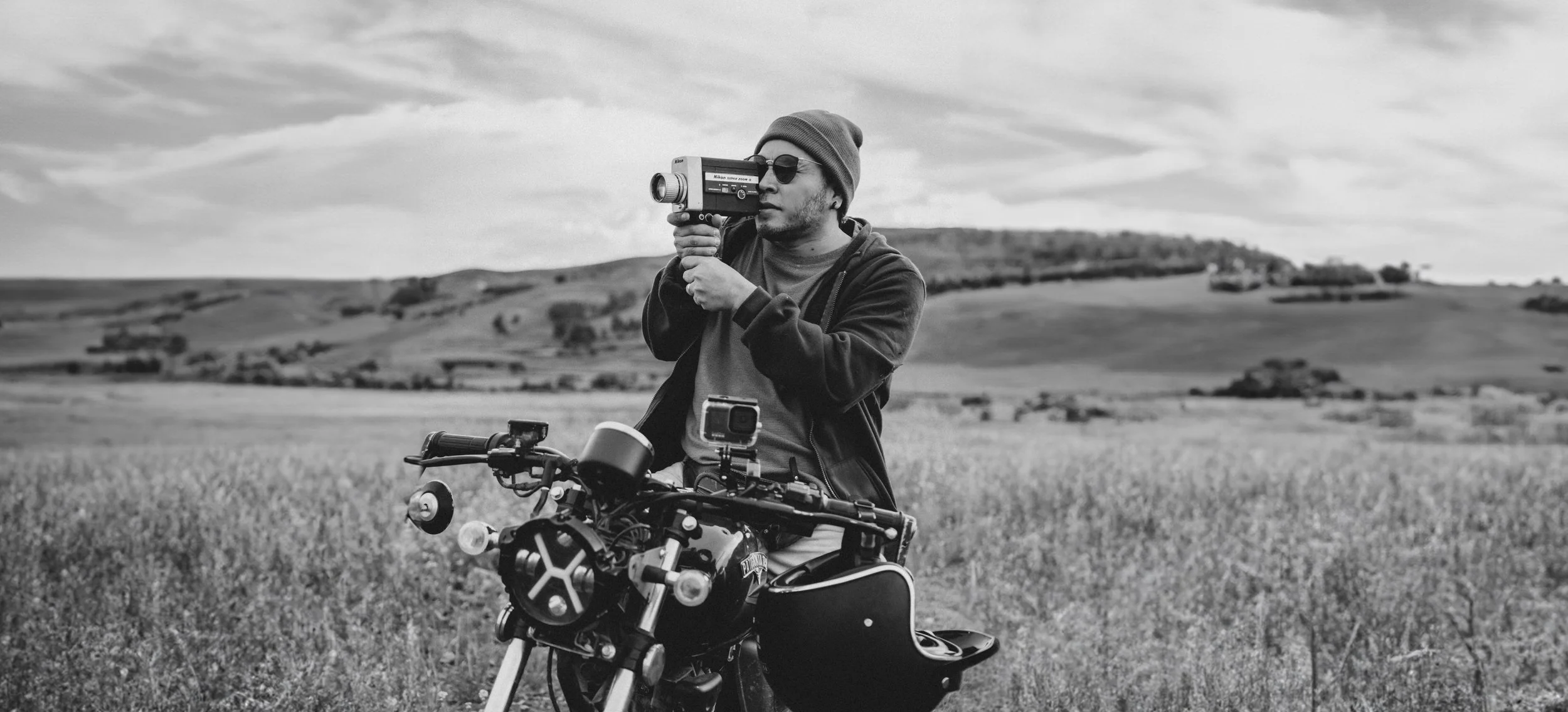 Un hombre con gorro y gafas de sol sostiene una cámara antigua en un campo abierto, junto a una motocicleta con una cámara GoPro en el manillar, en un paisaje rural con colinas y cielo nublado.