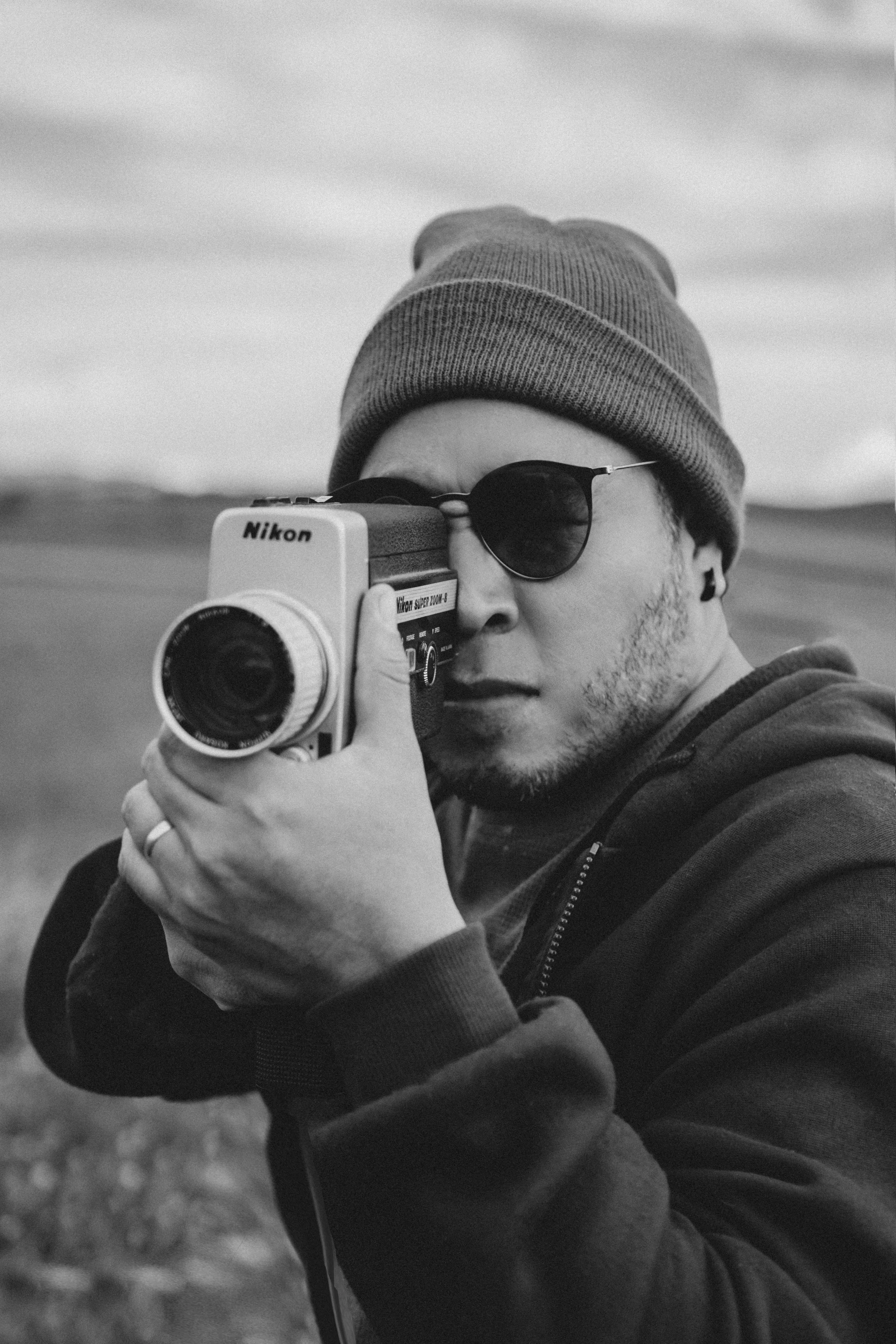 Joven sosteniendo una cámara Nikon, usando gafas oscuras, gorro y sudadera, en un paisaje al aire libre.