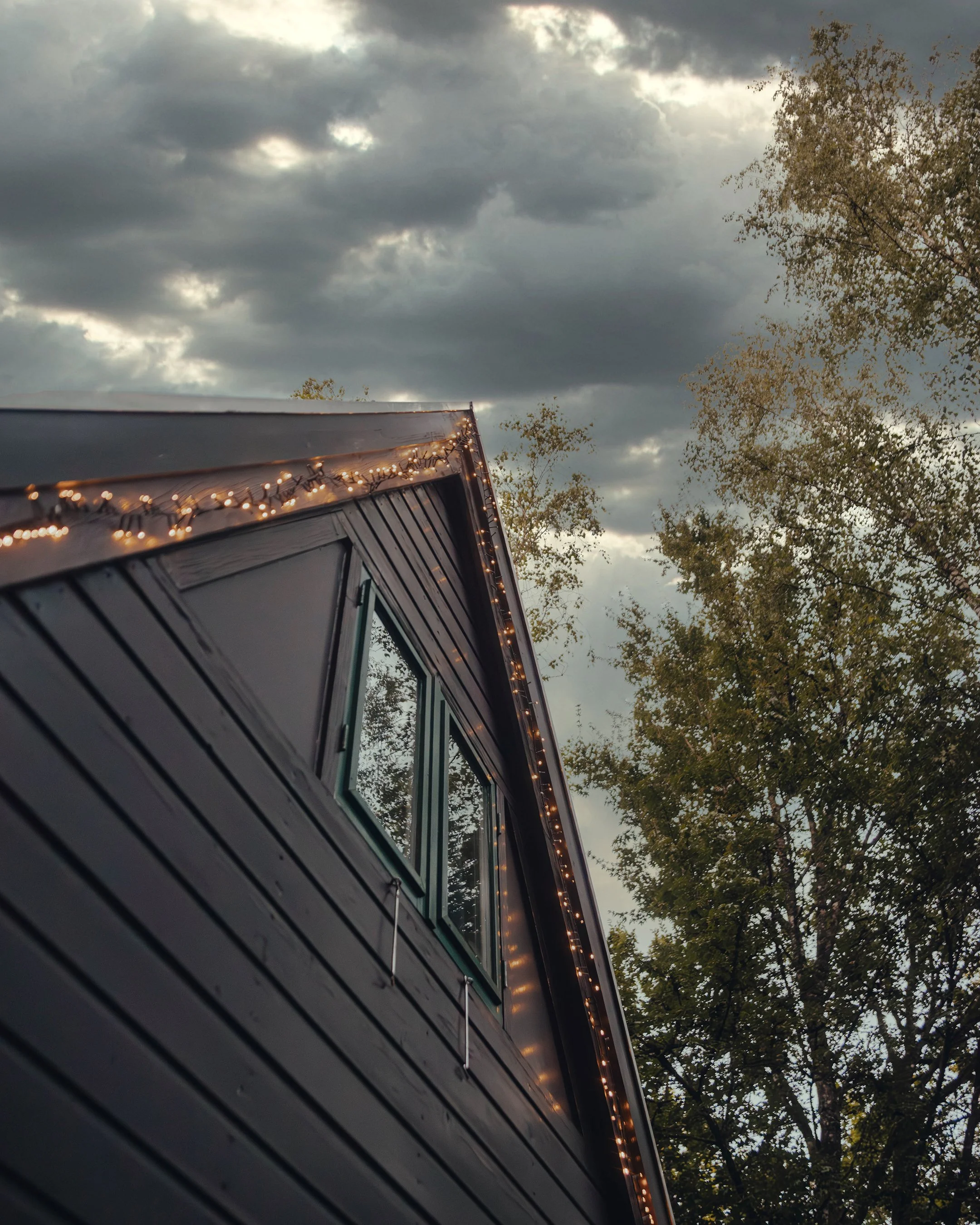 Close-up of a house's dark exterior with a window, decorated with string lights along the roof's edge. Tree with green leaves and a cloudy sky in the background.