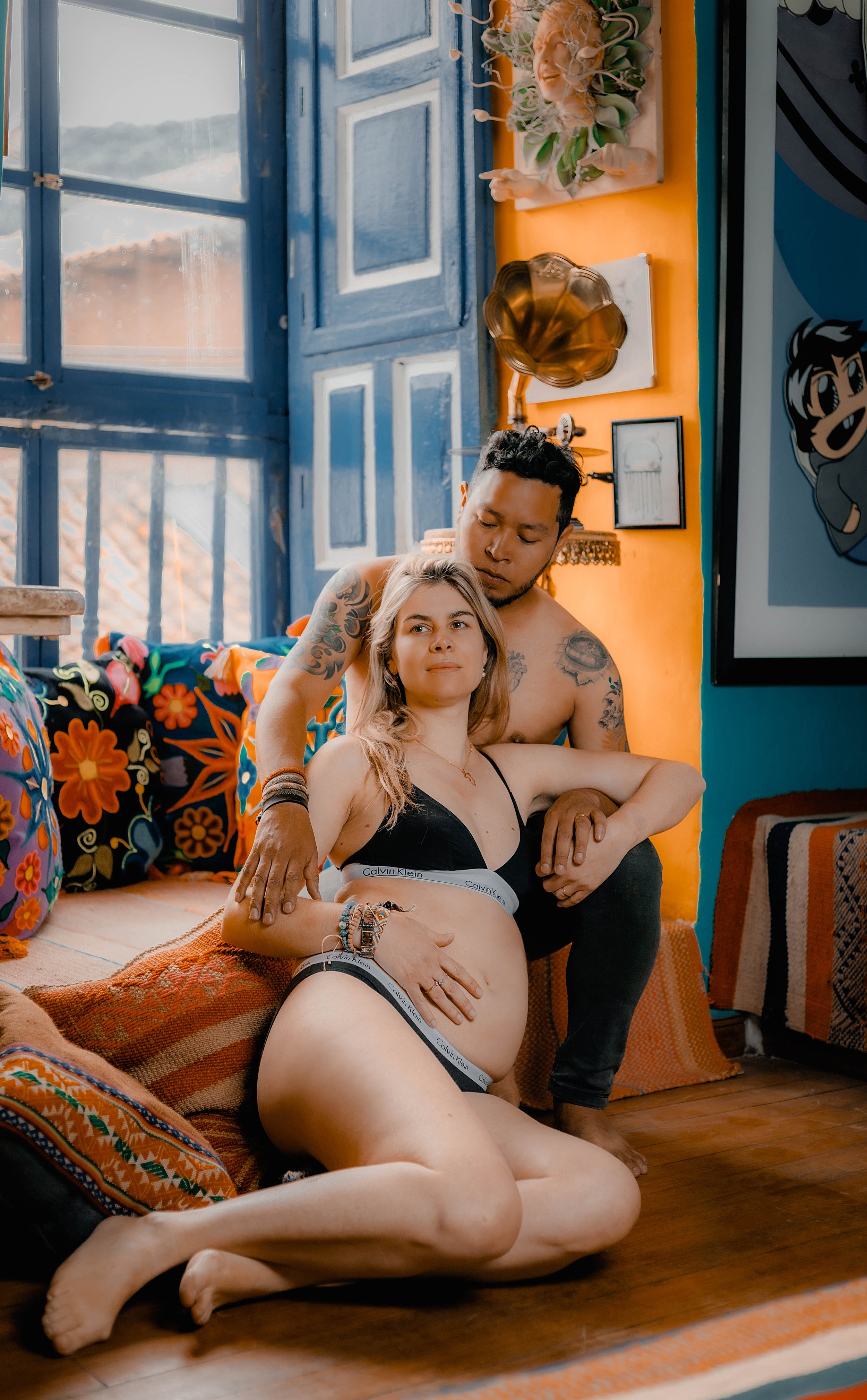 Una pareja en una habitación colorida, con la mujer embarazada sentada en el suelo y el hombre apoyándose en ella, rodeados de decoración vibrante y ventanas azules.