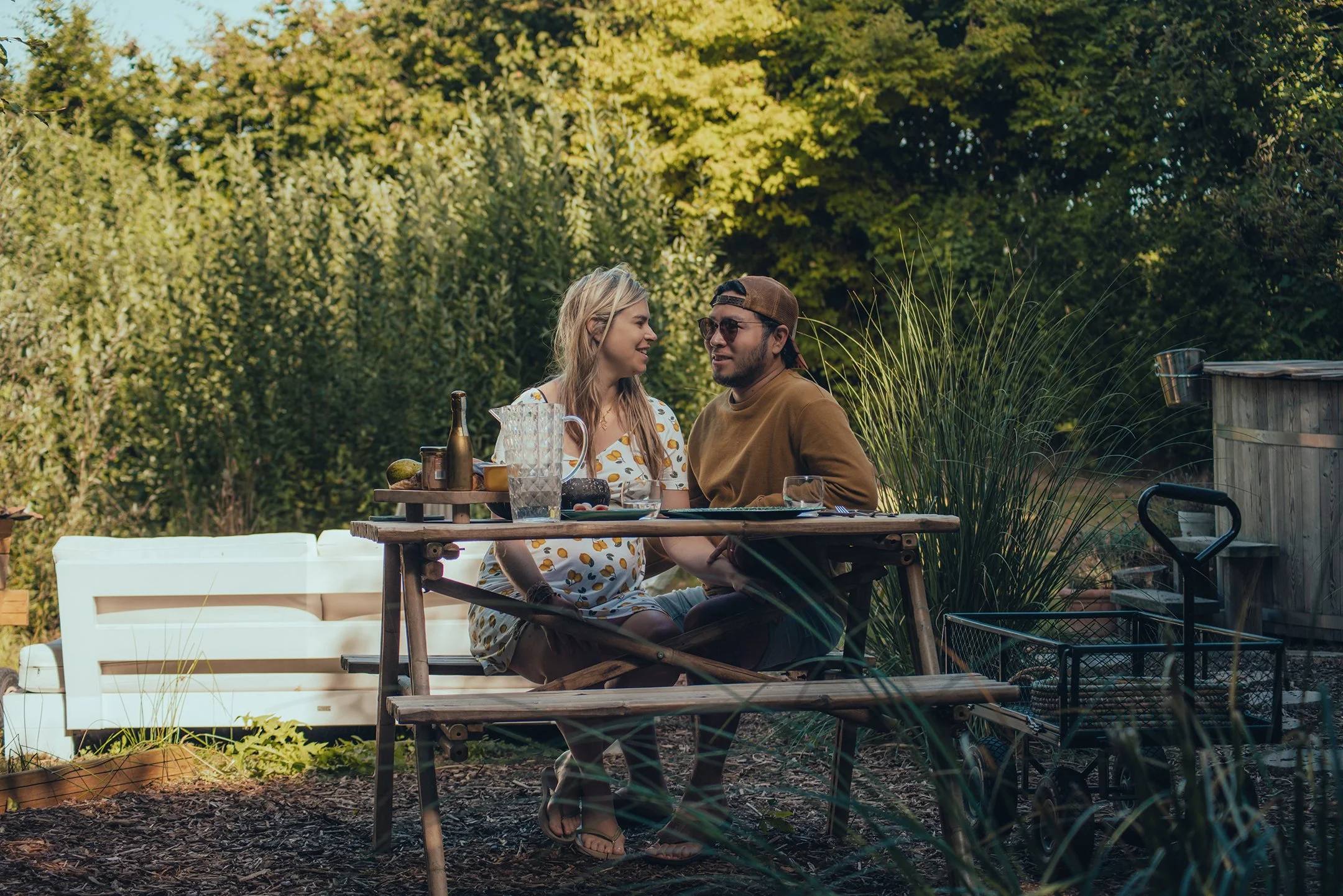 A couple sitting at an outdoor table in a backyard garden, sharing a moment. The table has a pitcher, bottles, and food, with lush green plants and trees in the background.