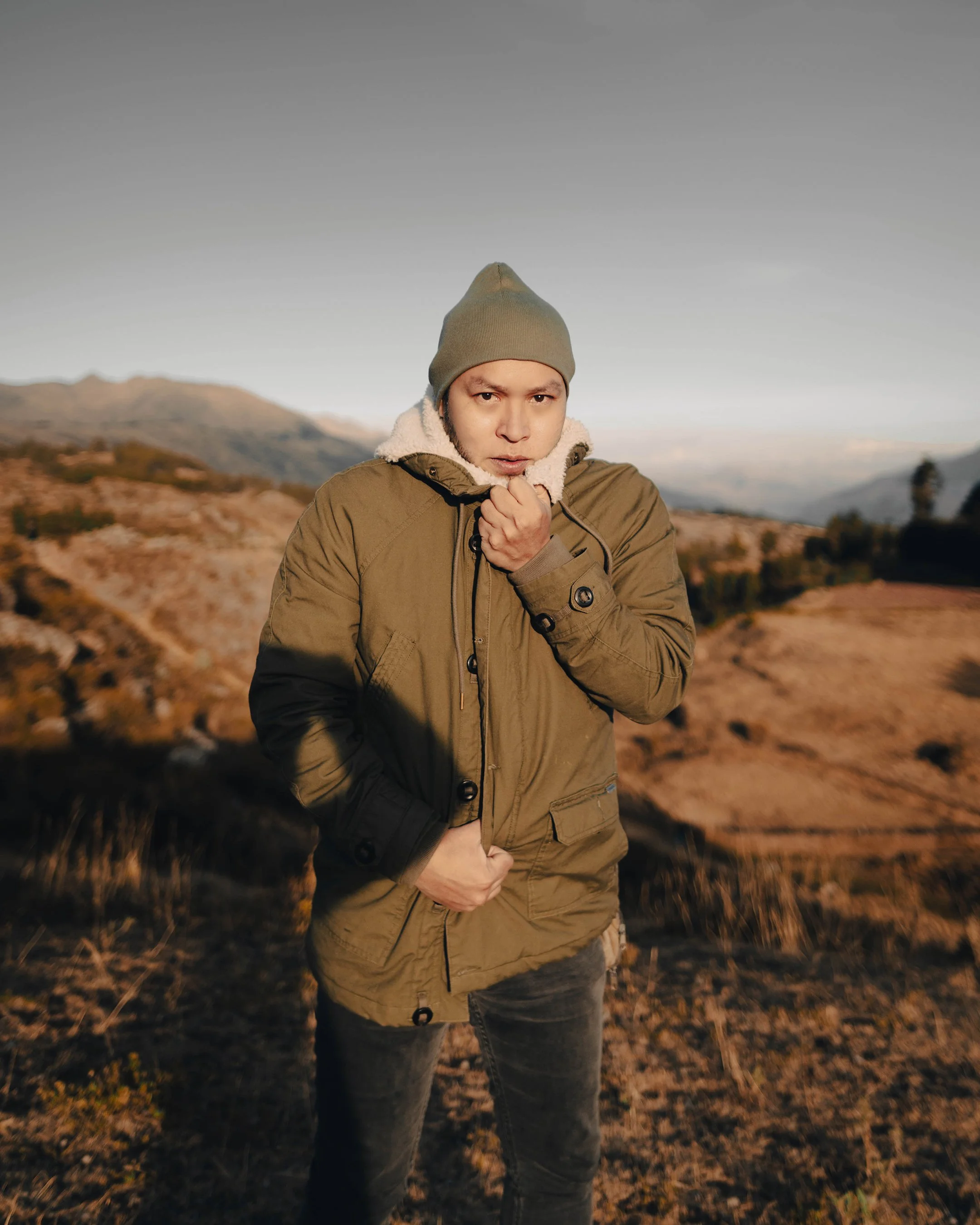 A young man in a green jacket and gray beanie stands outdoors in a mountainous landscape, holding the collar of his jacket near his chin, with a serious expression.
