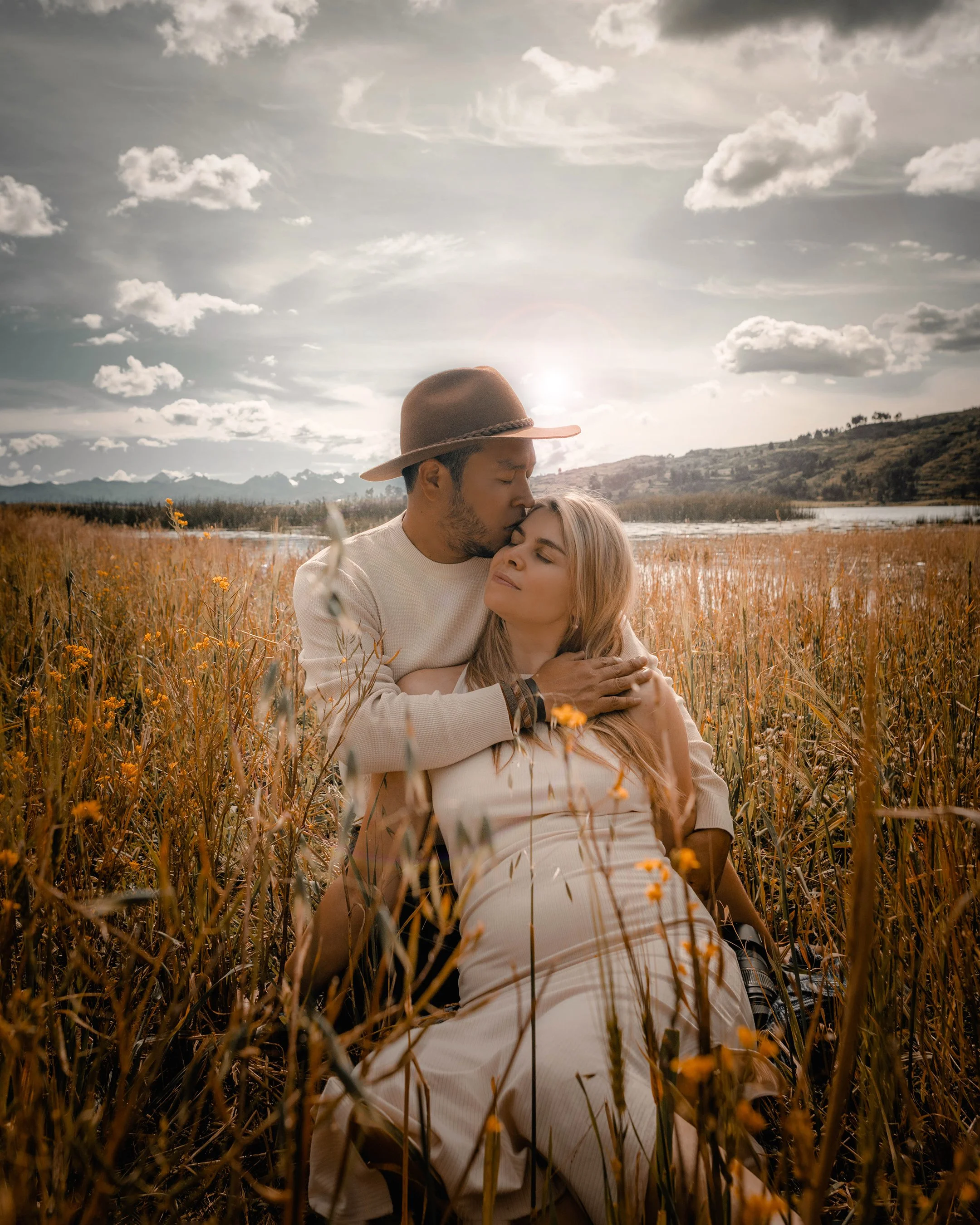 Una pareja en un campo abierto durante el atardecer, la mujer embarazada recibe un beso en la frente por parte del hombre, ambos parecen en un momento de ternura y amor.