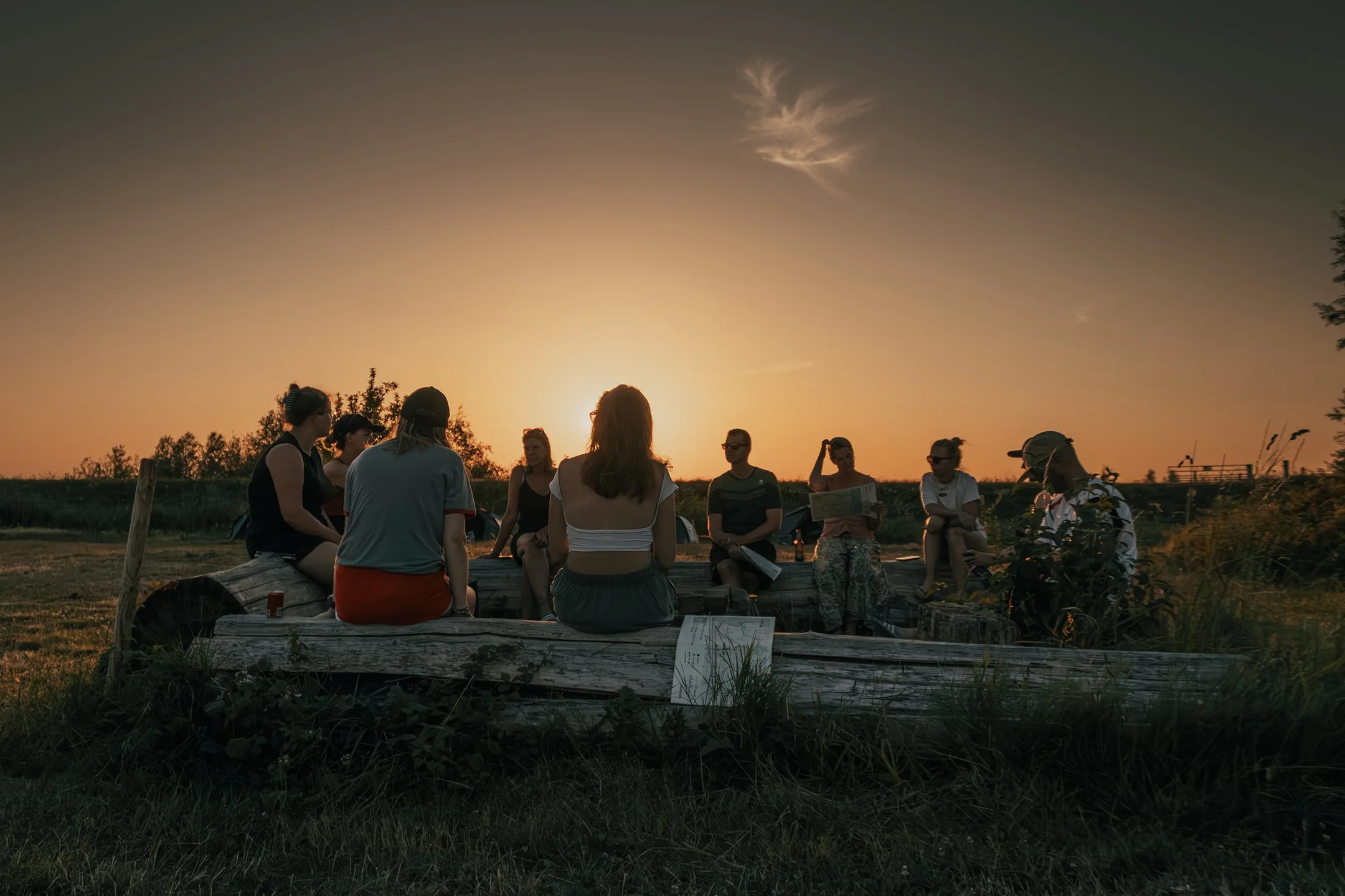 A group of people sitting on a log outdoors at sunset, engaged in conversation, with some reading and others wearing sunglasses.