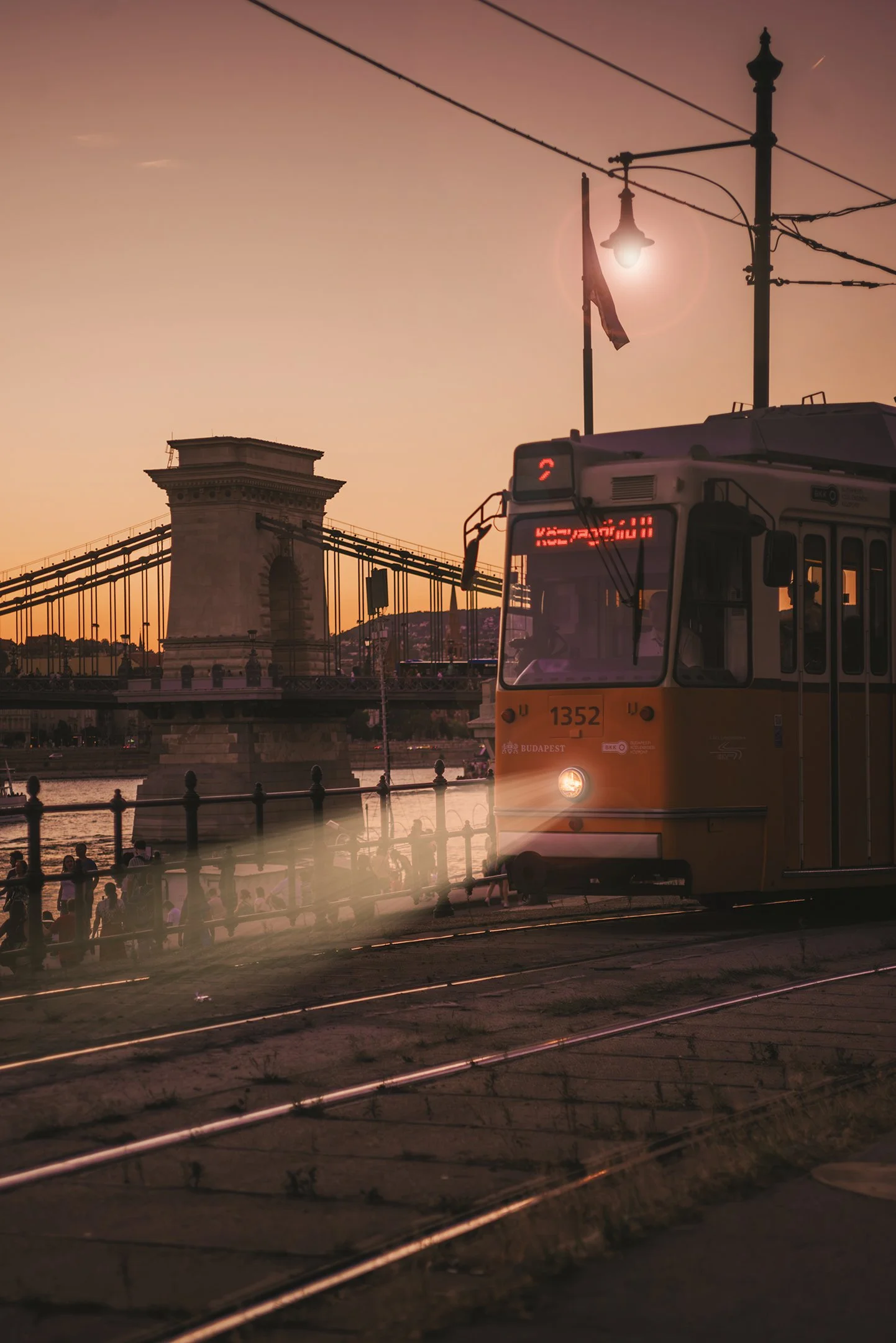 A tram moving on tracks near a river at sunset, with a bridge and people along the riverside in the background.