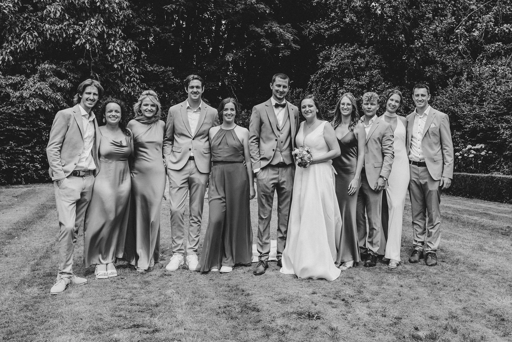Un grupo de personas en una celebración al aire libre, posiblemente una boda, con árboles y naturaleza de fondo, vestidos elegantemente, en tonos claros y oscuros.