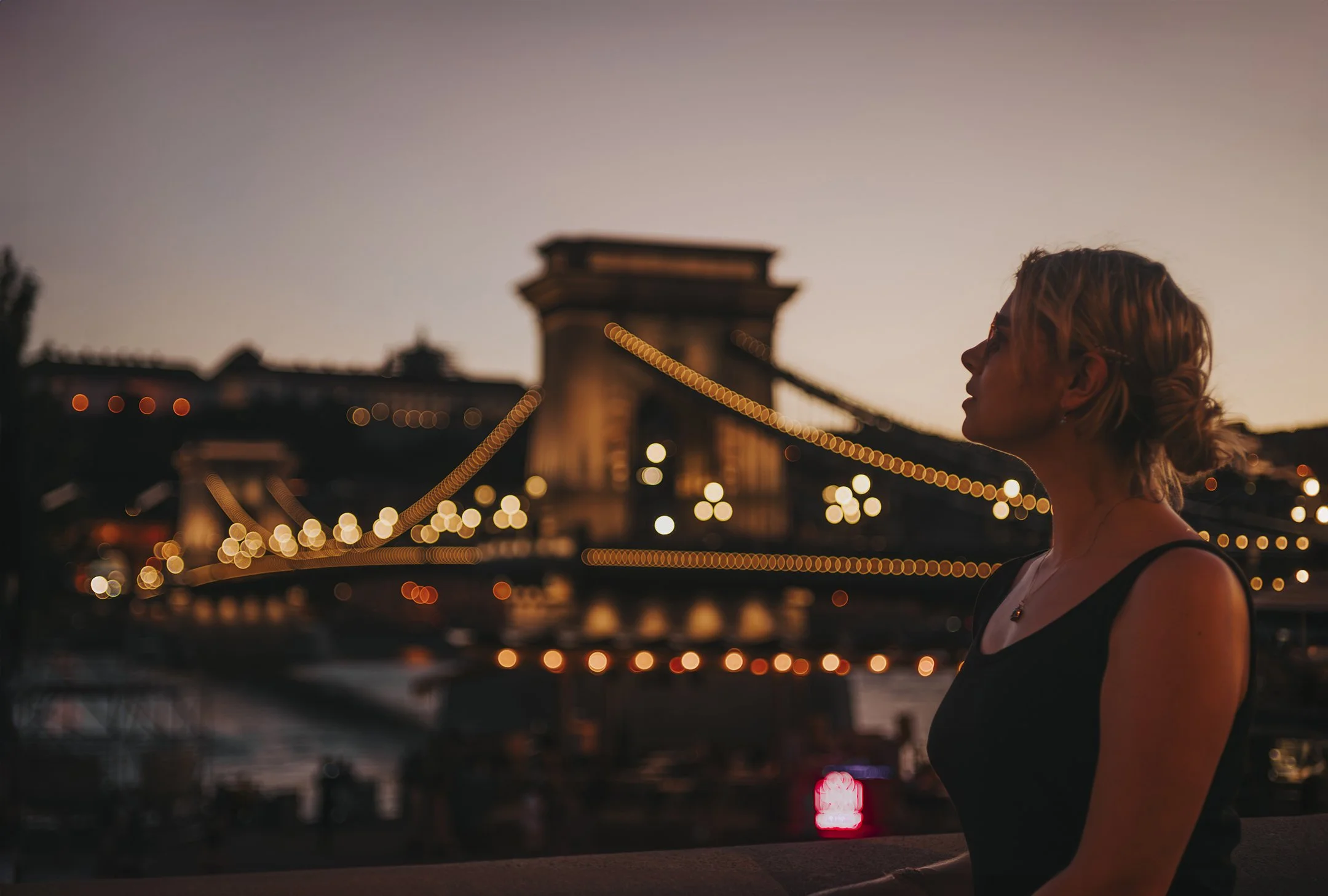 A woman with blonde hair in a loose bun, wearing a black dress, standing in profile against the backdrop of a lit-up bridge at dusk.