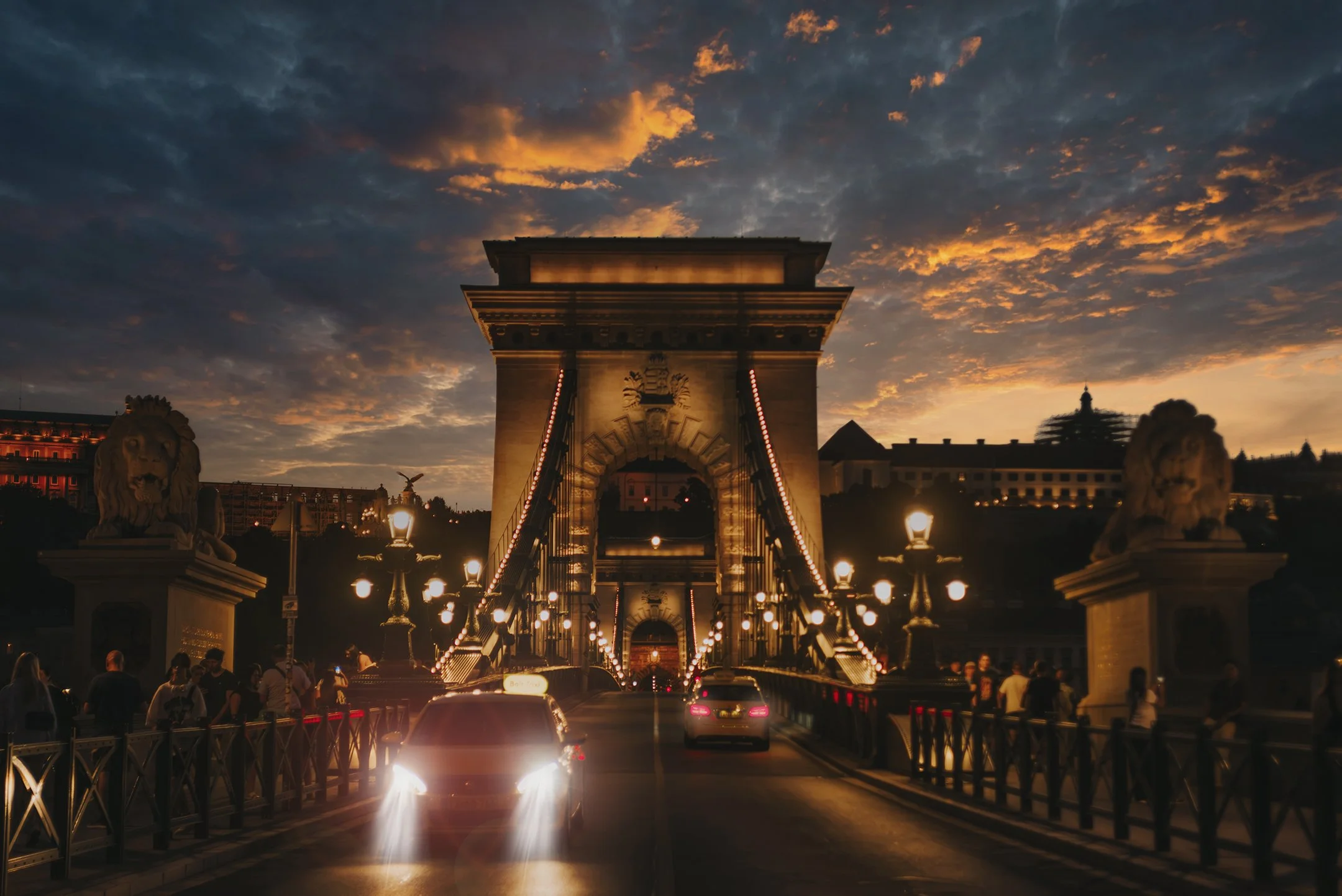 A suspension bridge illuminated with string lights at sunset, with cars driving across and people walking on the sidewalk, lion statues on the sides, under a partly cloudy sky.