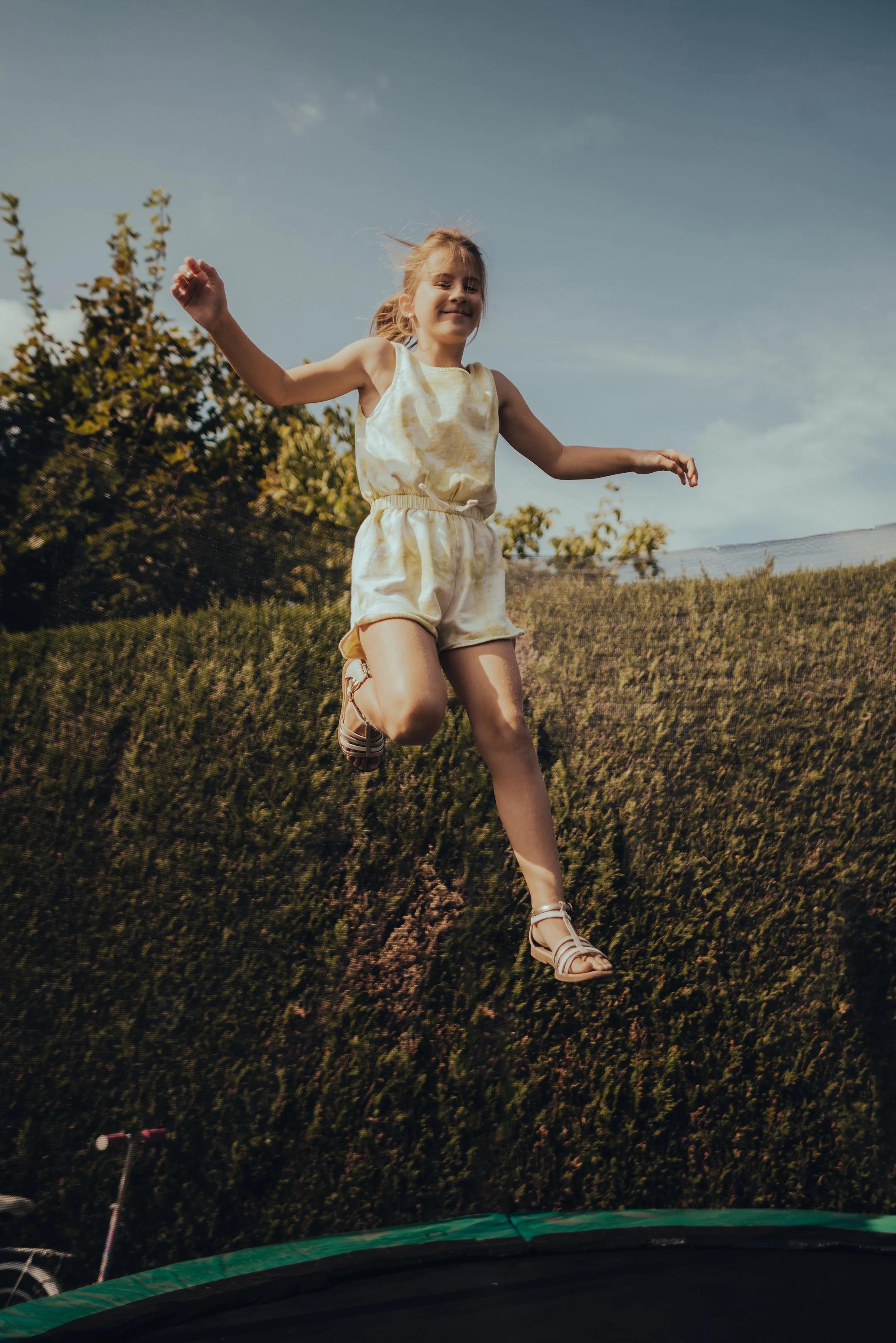 Girl jumping on a trampoline outdoors with a hillside and trees in the background.