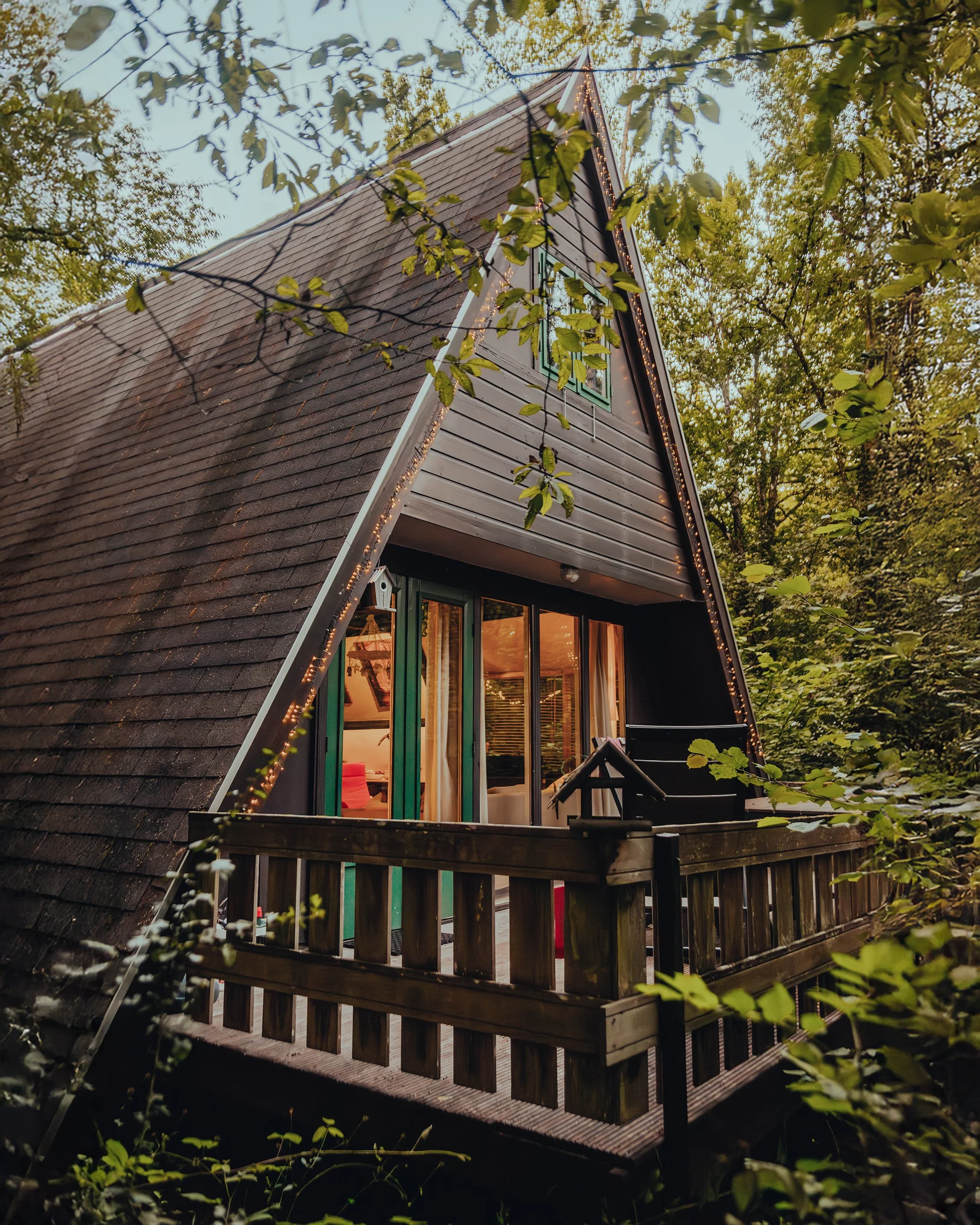 Aerial view of an A-frame cabin surrounded by trees with a small wooden balcony and warm lighting inside.