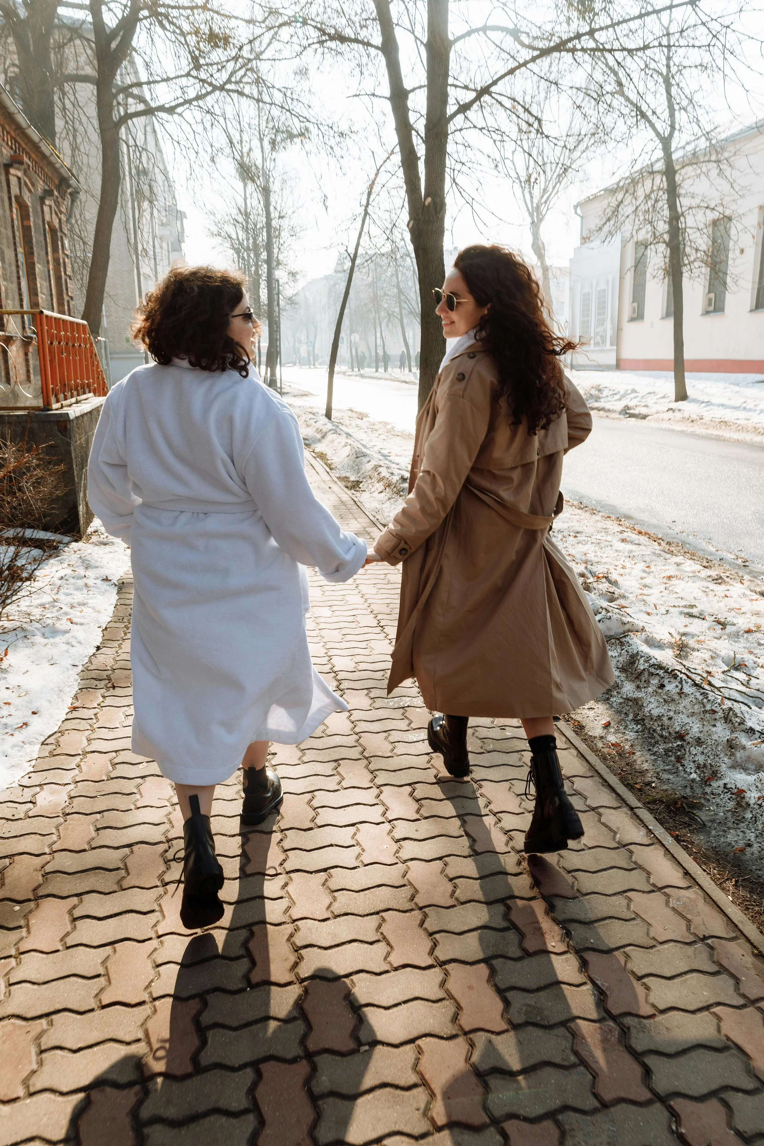 Dos mujeres caminando de la mano por la acera en un día soleado de invierno, vestidas con abrigos y botas negras, con árboles desnudos a su alrededor.