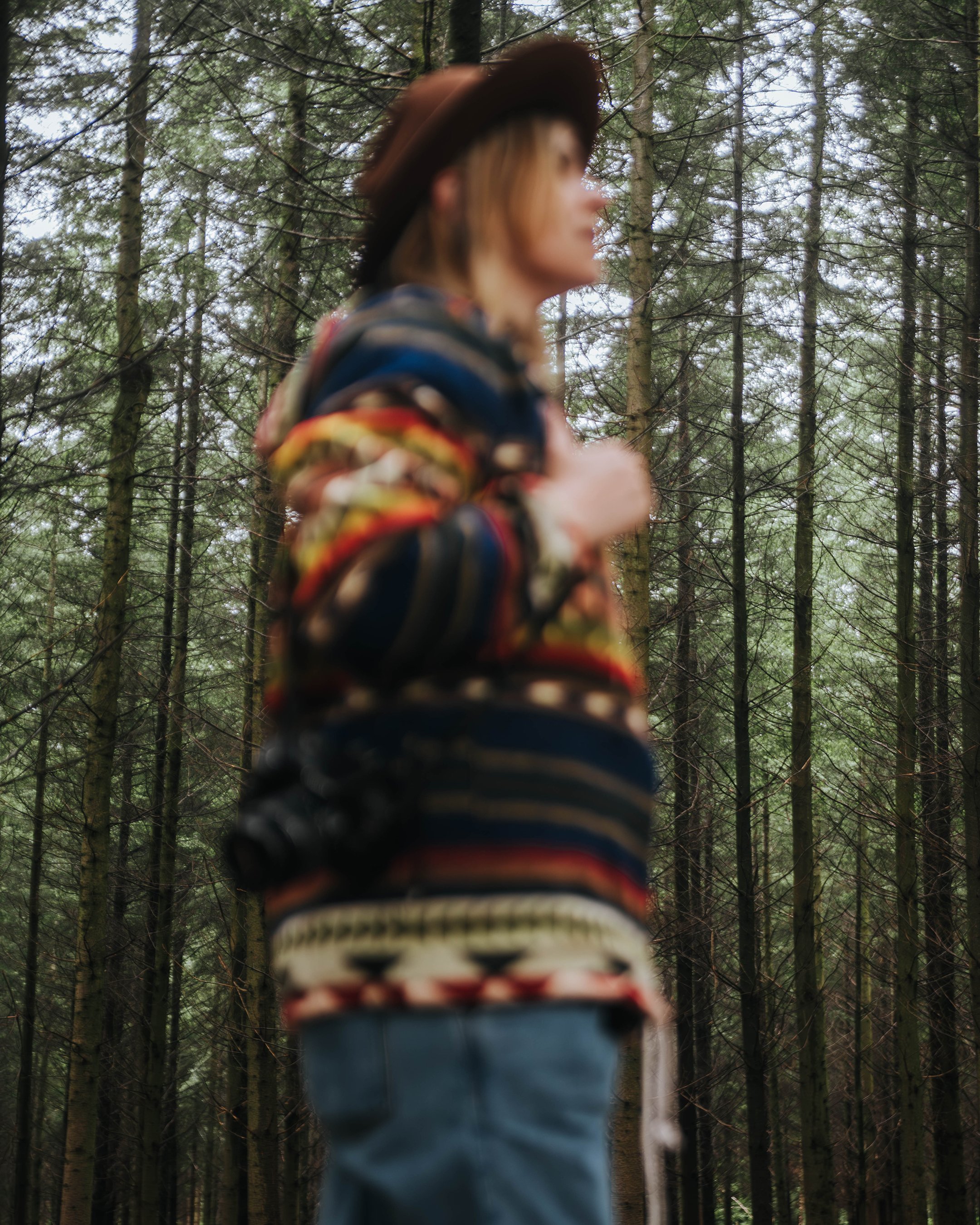 Blurred image of a woman with a wide-brimmed hat in a forest, wearing a colorful patterned sweater and blue jeans.