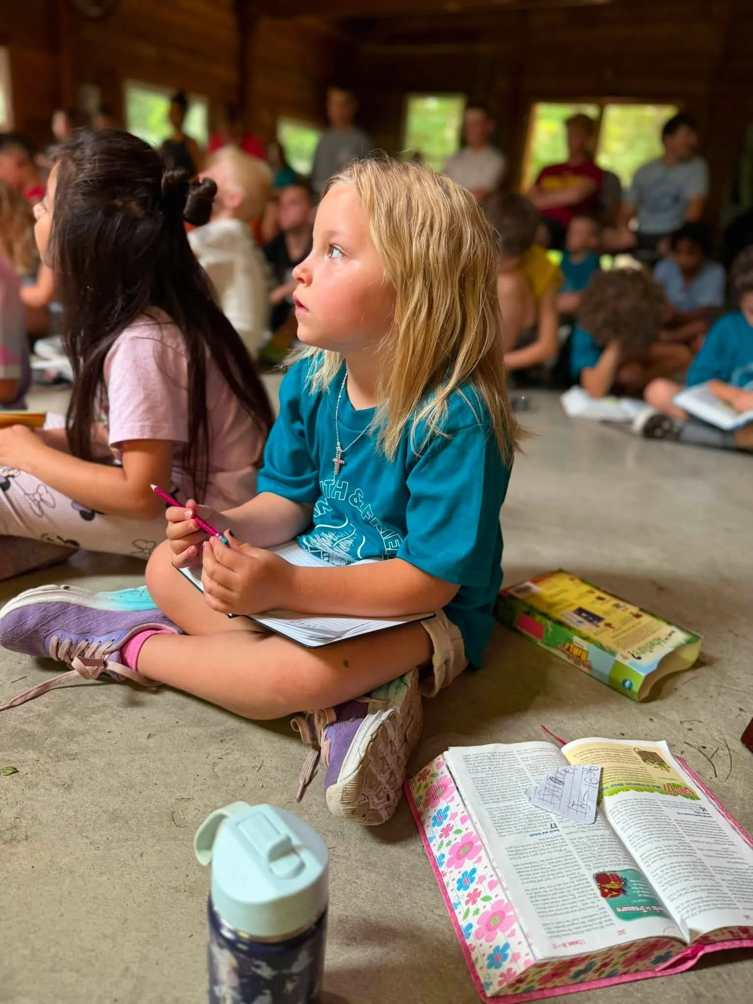 A young girl with blonde hair sitting on the floor in a room with wooden walls, surrounded by open books and school supplies. Many other children are visible in the background, some sitting and some standing, inside a rustic cabin or classroom with windows showing greenery outside.