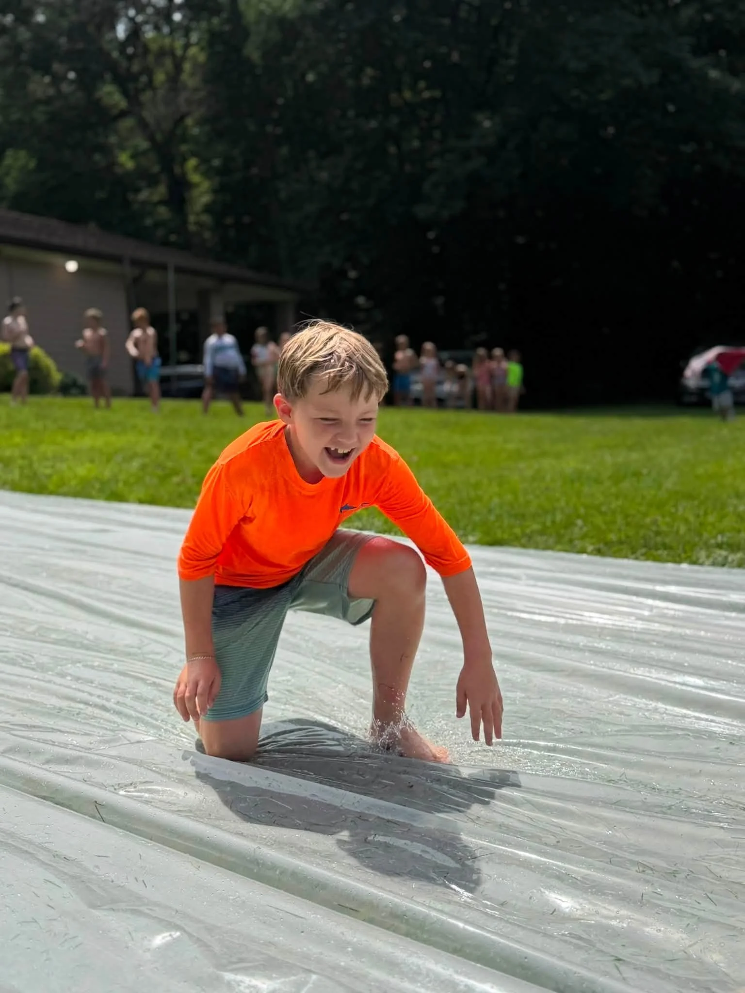 A young boy in an orange shirt and gray shorts is kneeling on a slip-and-slide in a backyard, smiling and playing in water amid other children and adults in the background.