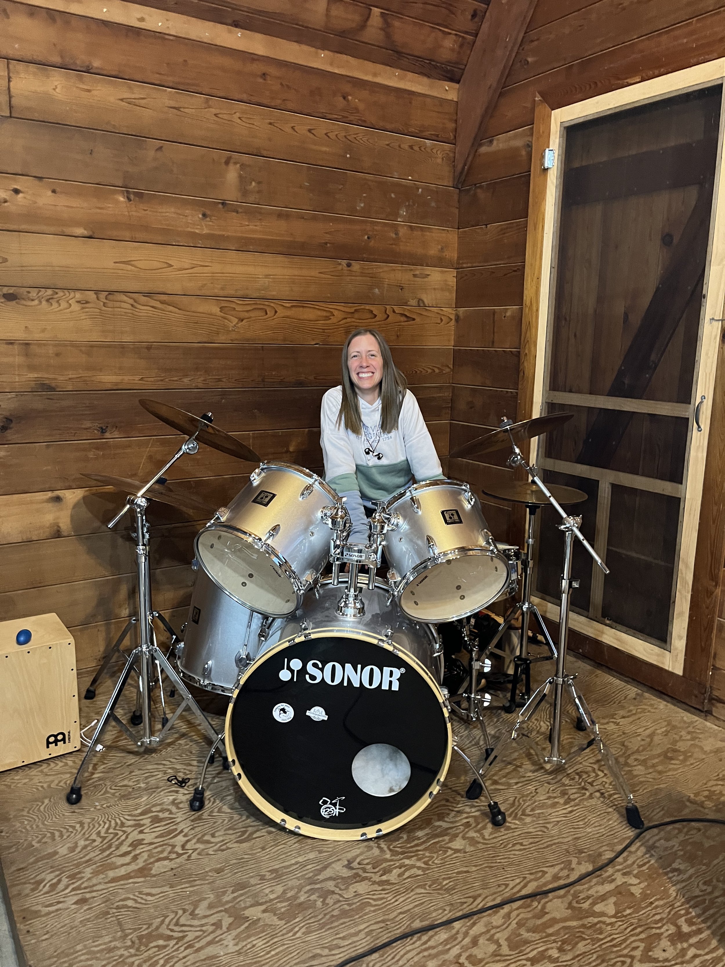 A smiling woman sitting behind a drum set with a wooden wall background, a window, and a cajón drum on the floor.