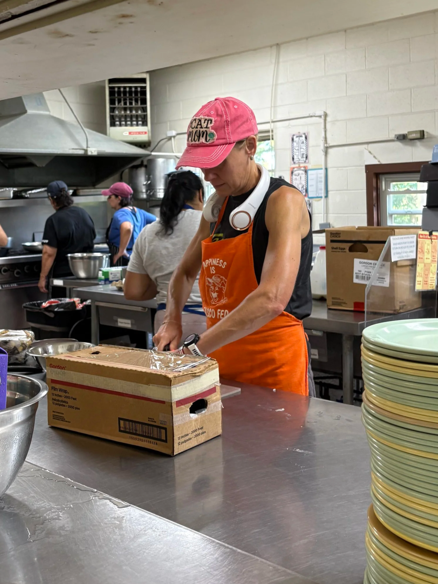A woman wearing a pink cap with 'CAT MOM' written on it, an orange apron, and headphones around her neck working in a commercial kitchen, with other people in the background preparing food.