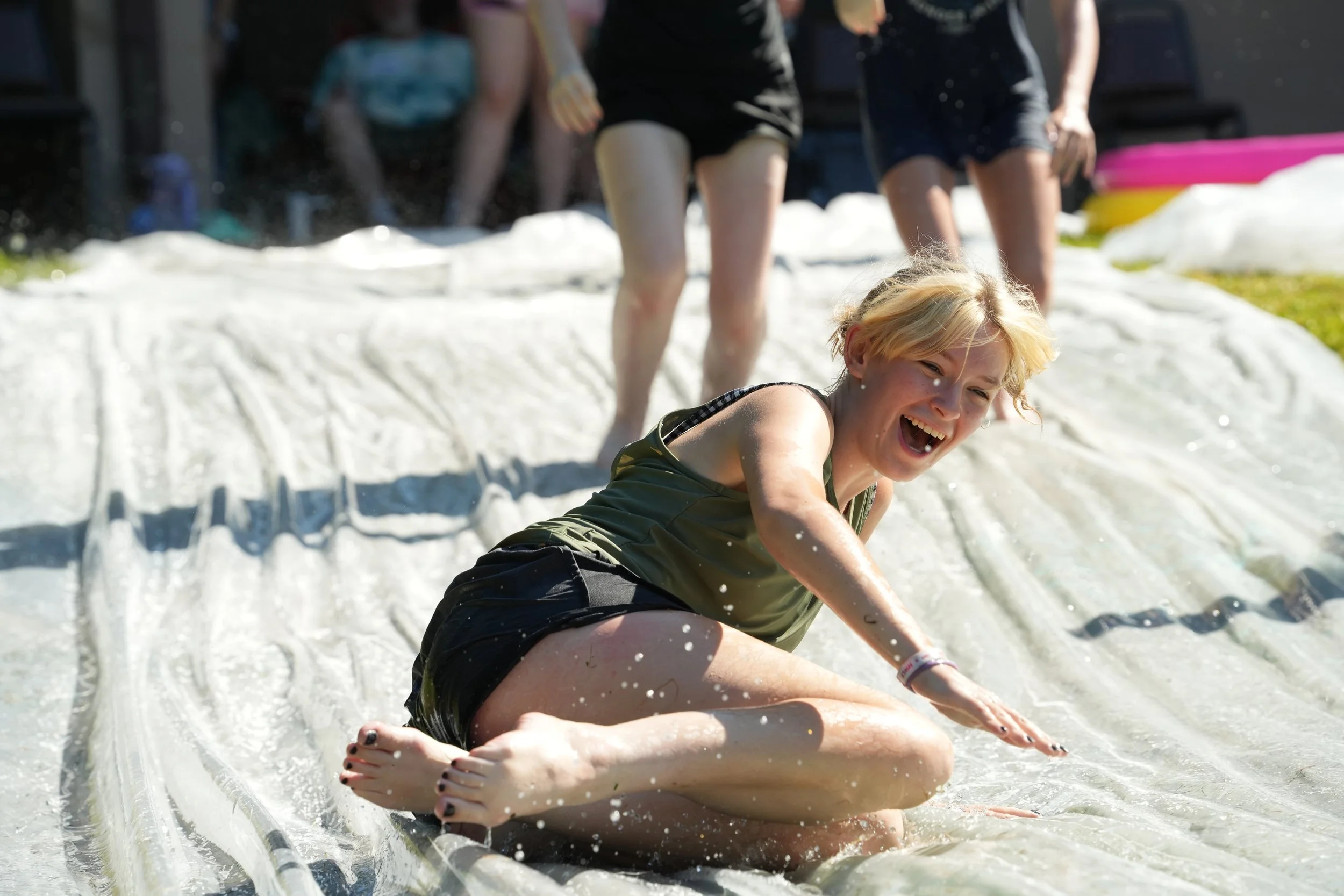 A young woman with short blonde hair lying on a slip and slide in a backyard, laughing and enjoying a water activity on a sunny day.