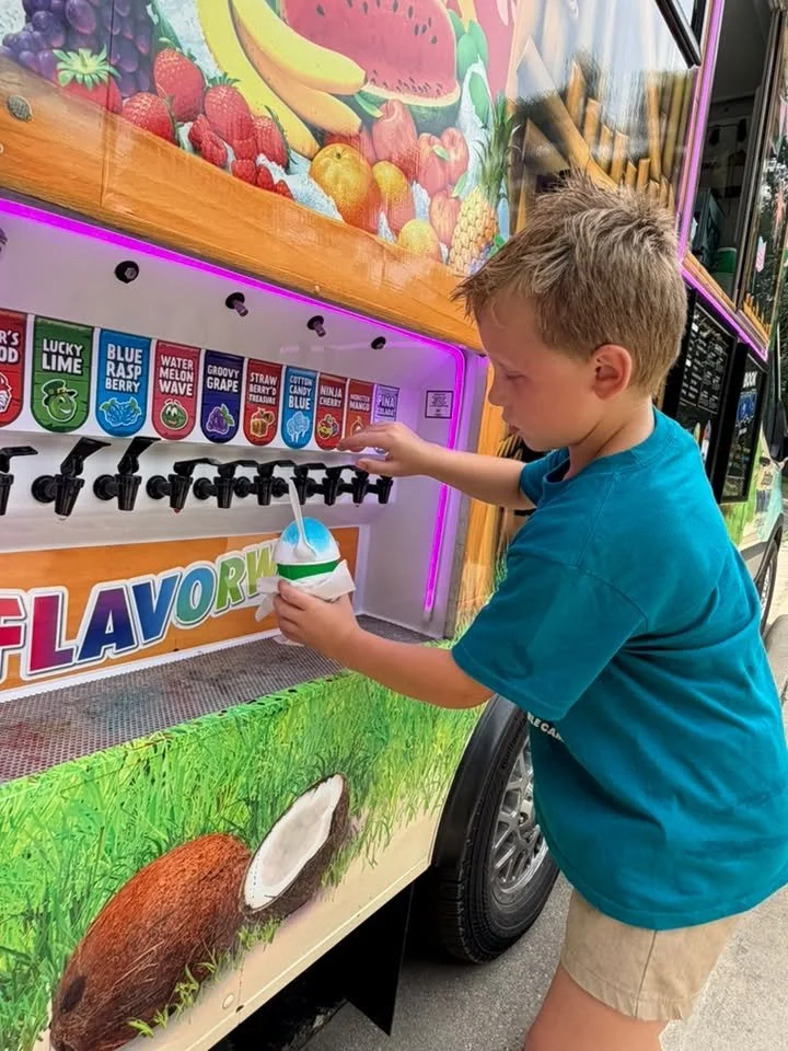 A young boy in a blue shirt and tan shorts is at a colorful ice cream or slush machine outside, filling a cup with a blue and white striped straw. The machine has various flavors displayed with labels, and the background features artwork of fruits such as bananas, strawberries, and watermelon.