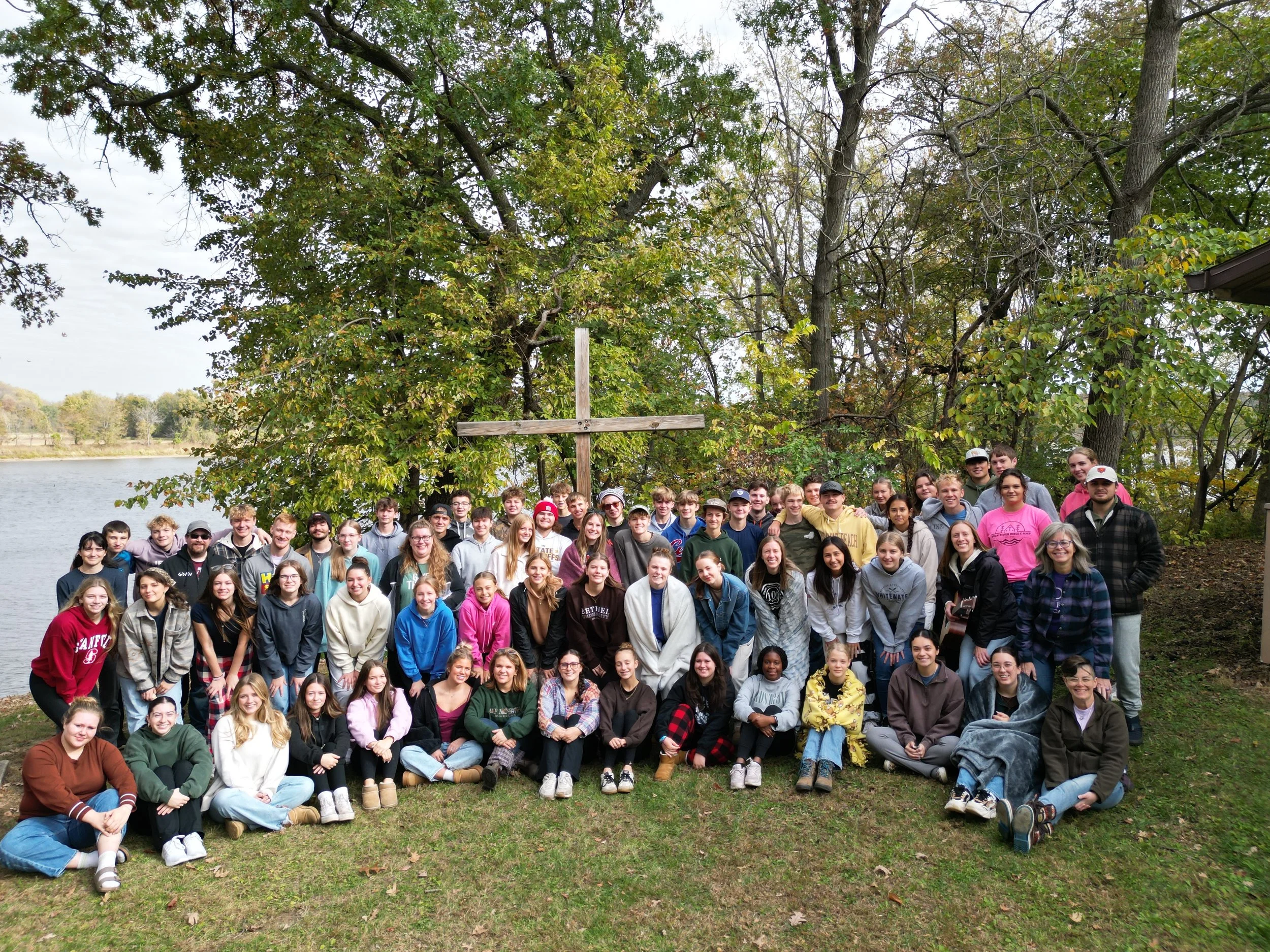 A large group of young people, mostly teenagers, gathered outdoors on grass near a lake, posing for a group photo in front of tall trees with a wooden cross among the trees in the background.