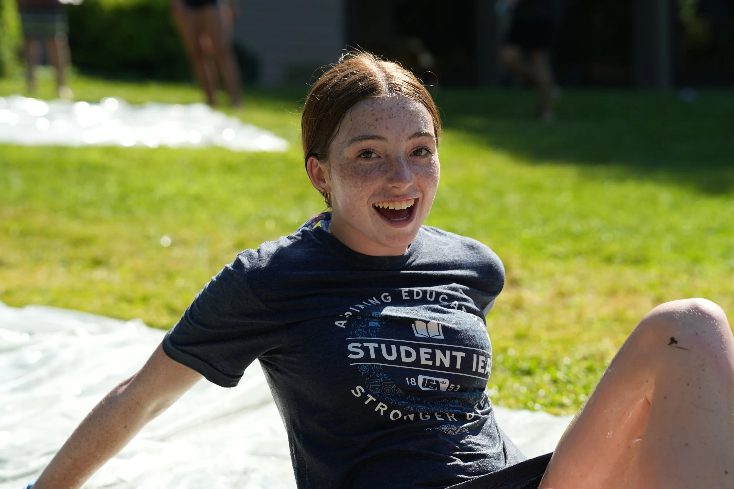 A young girl with freckles and brown hair smiling outdoors on a sunny day. She is wearing a dark t-shirt with white text and illustrations, and is sitting on grass with blurred trees and a pond in the background.