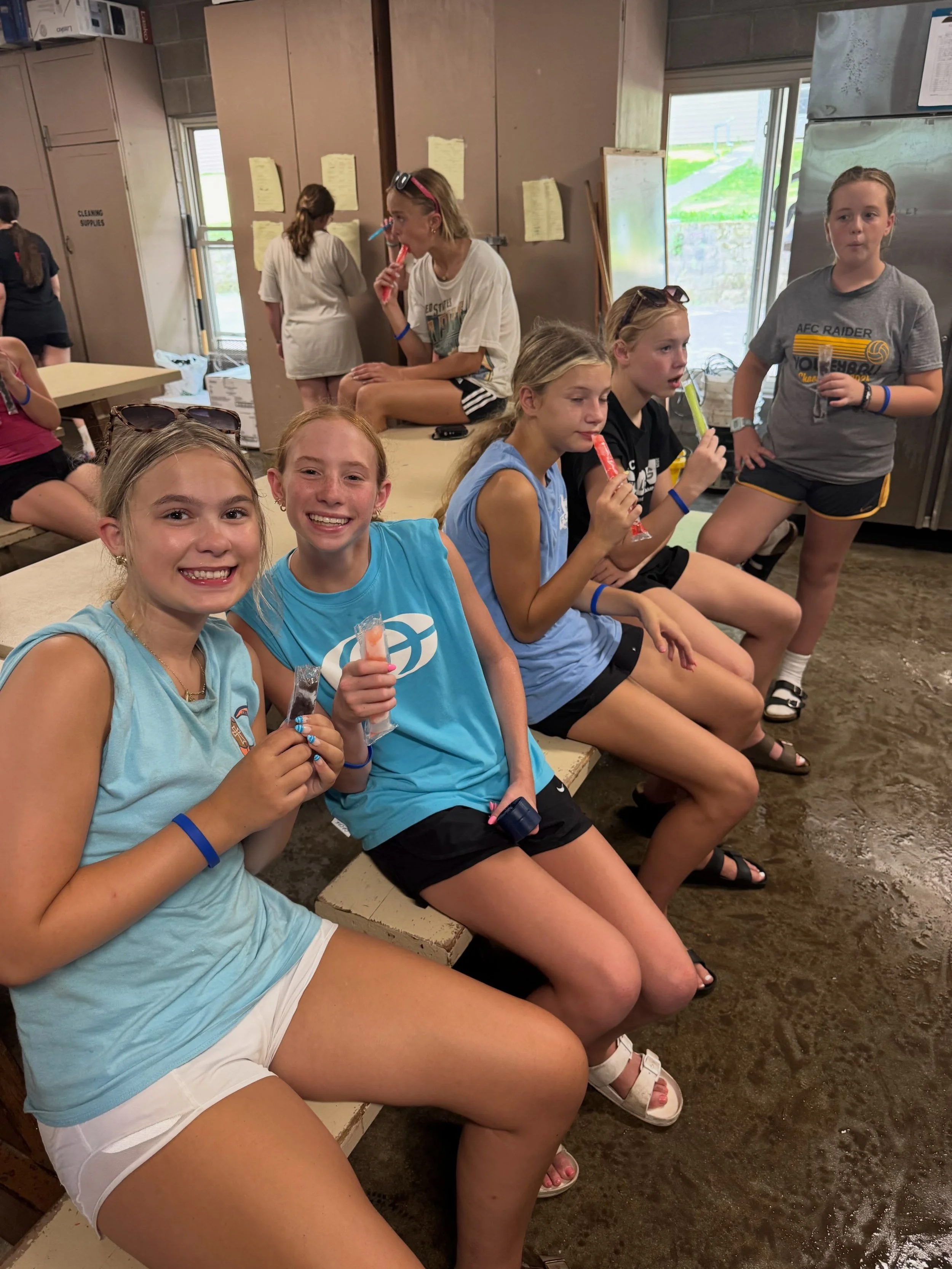 Group of young girls sitting and standing indoors, holding popsicles, smiling, and chatting.