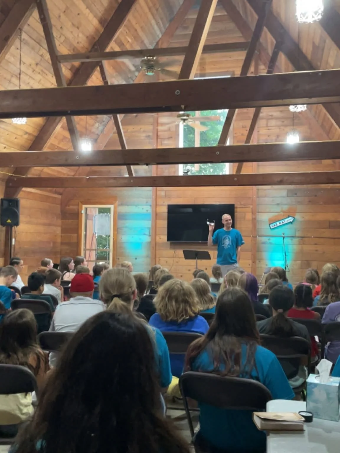 A speaker in a blue shirt standing at the front of a wooden beamed hall, addressing a large audience of children, with a TV mounted on the wall behind him and a sign on the right.