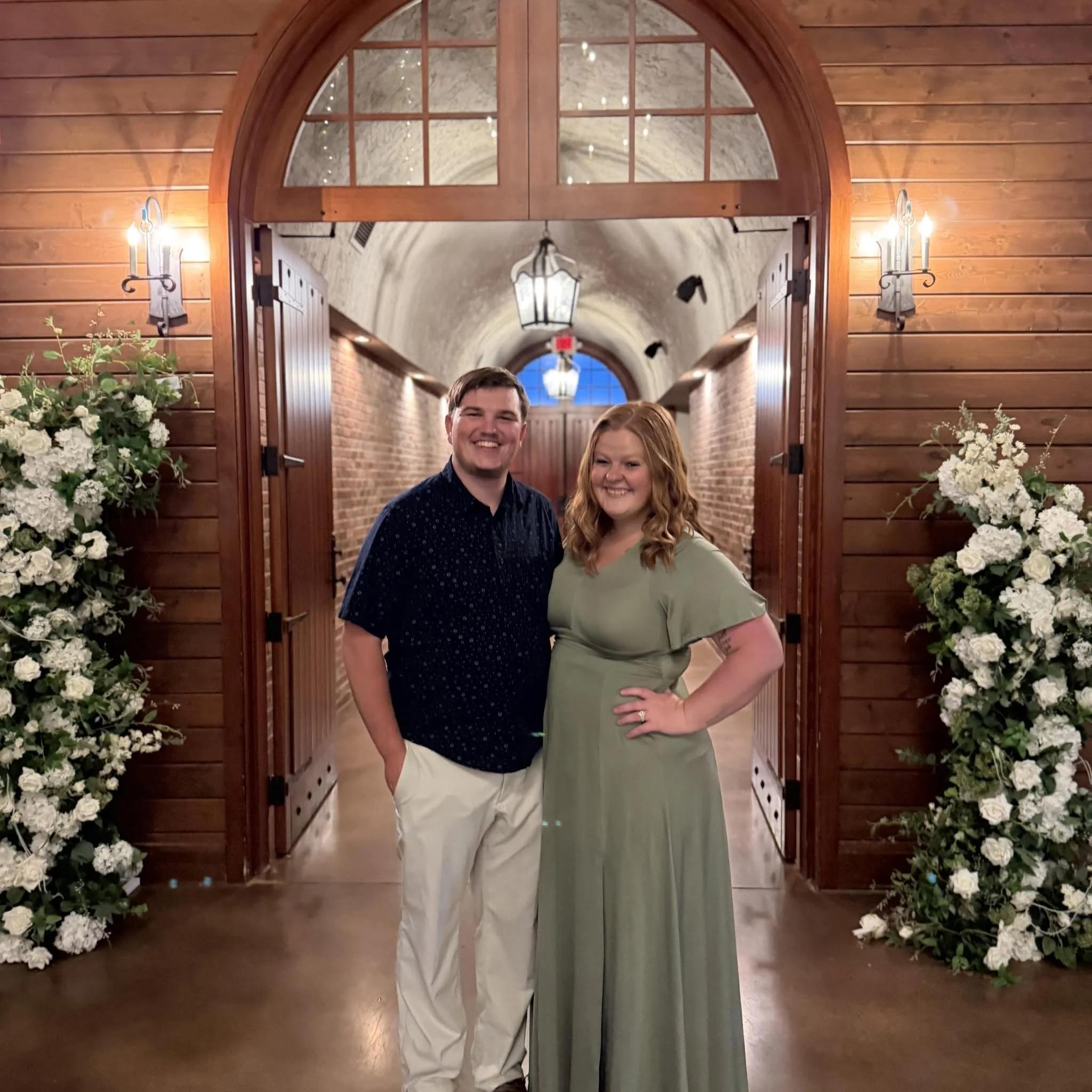 A happy man and woman standing in front of a wooden arch doorway decorated with white flowers, inside a venue with brick walls and lantern-style ceiling lights.