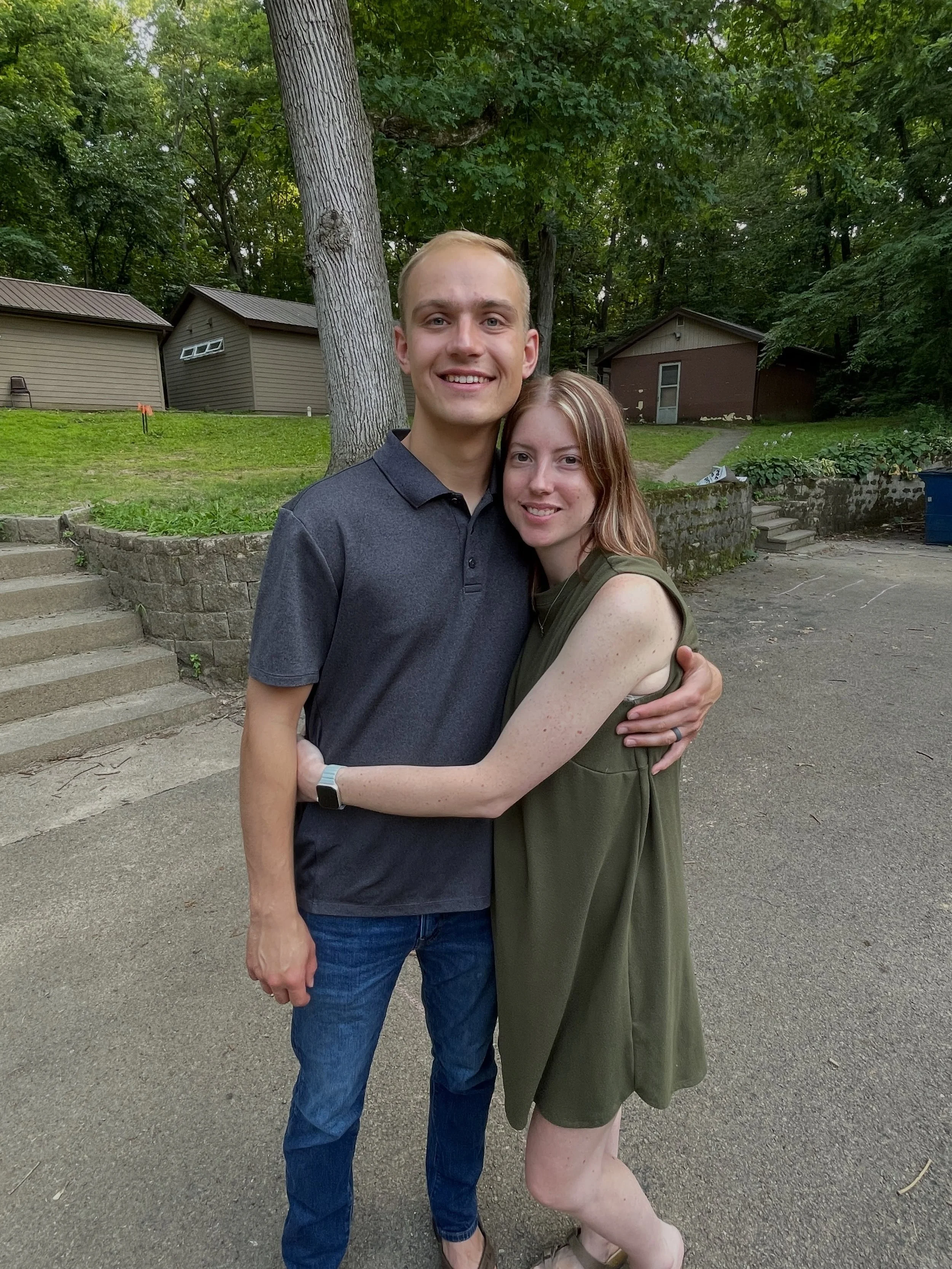 A young man and woman hugging outdoors near a tree and small houses, smiling at the camera.