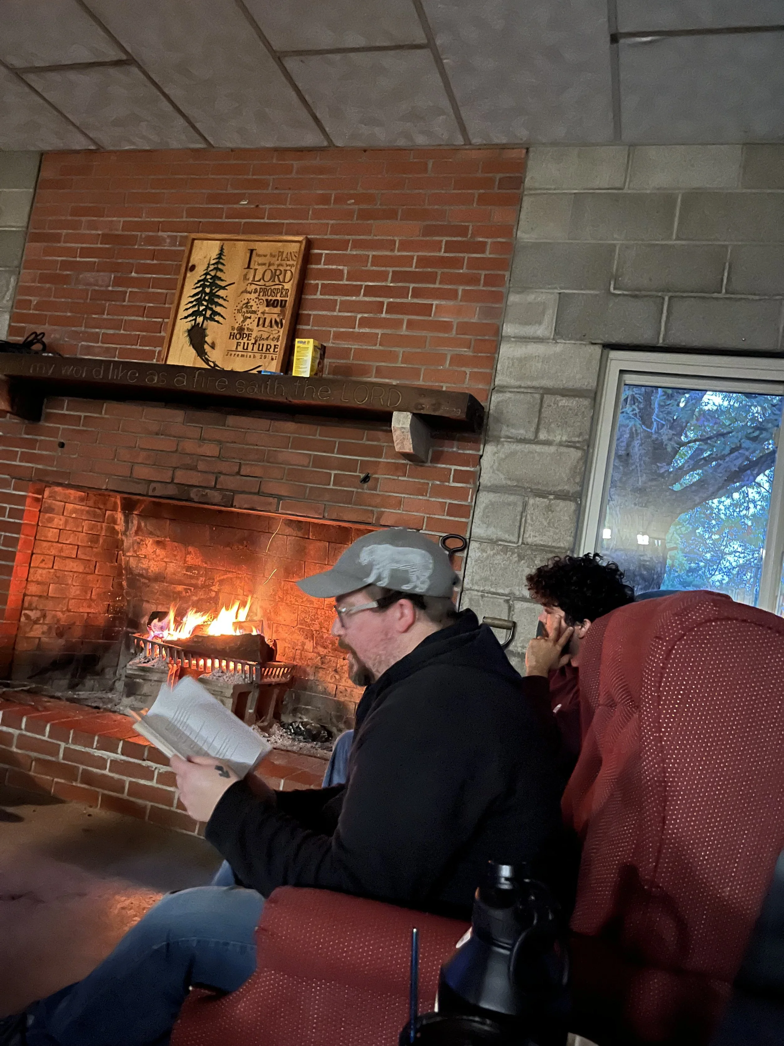 People sitting in front of a fireplace with a brick chimney, reading books. One person is wearing a gray cap, and there is a window showing trees outside.