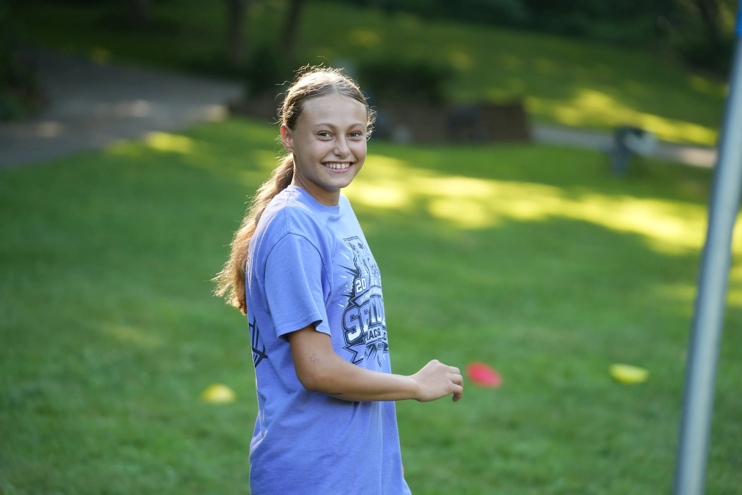 A young girl with long blonde hair in a ponytail smiling outdoors on a sunny day, wearing a blue T-shirt with a graphic design, standing in a grassy park with trees in the background.