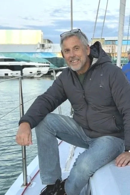 A man sits on the edge of a boat, smiling, with marina and boats in the background.