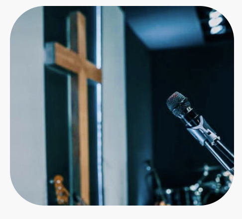 A microphone on a stand in a stage or church setting with a wooden cross on the wall behind it.