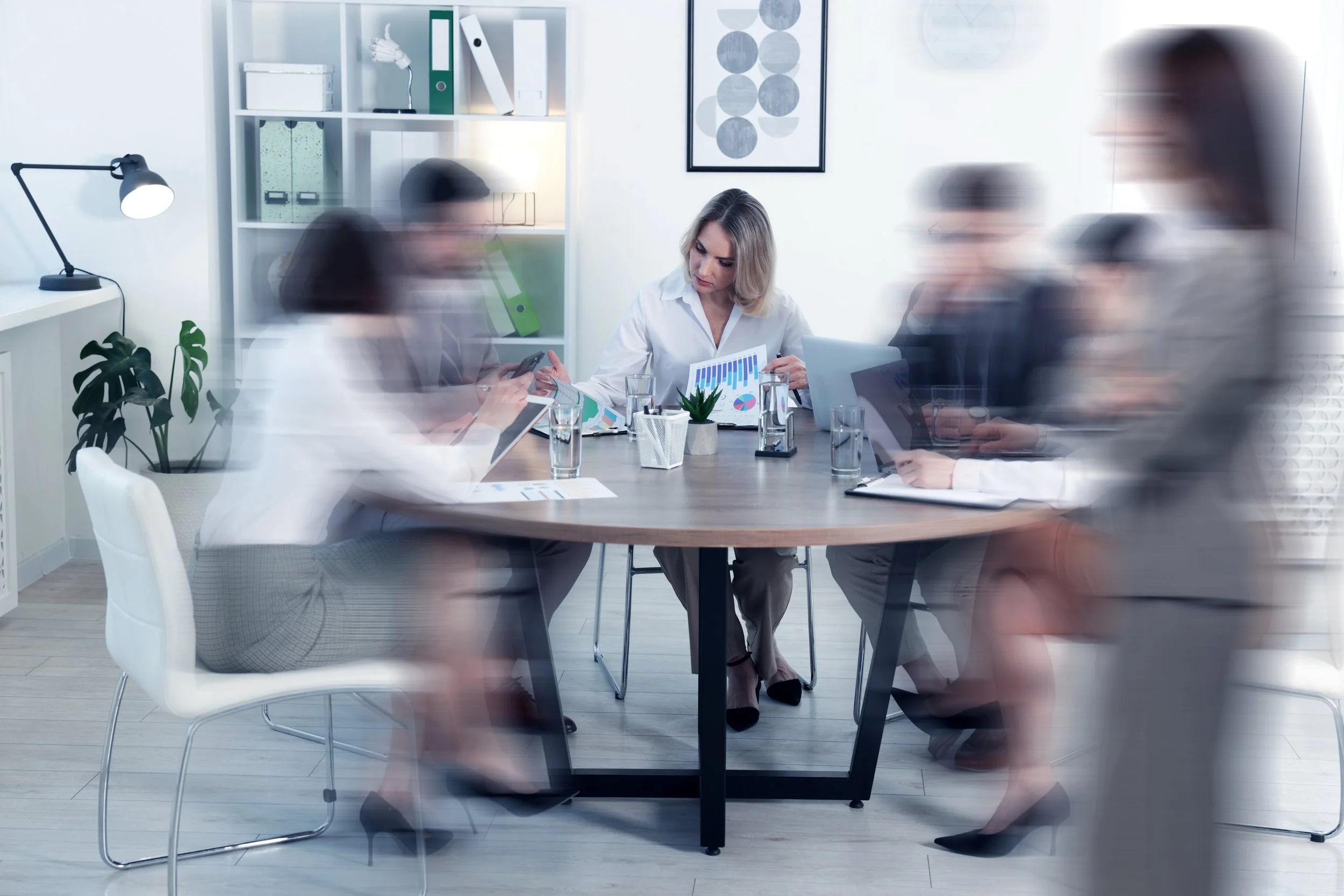 A business meeting room with six blurred-out people walking past or standing, and a woman sitting at a table reviewing charts and graphs. The room has white walls, a bookshelf with binders, a desk lamp, a small potted plant, and a framed geometric art piece.