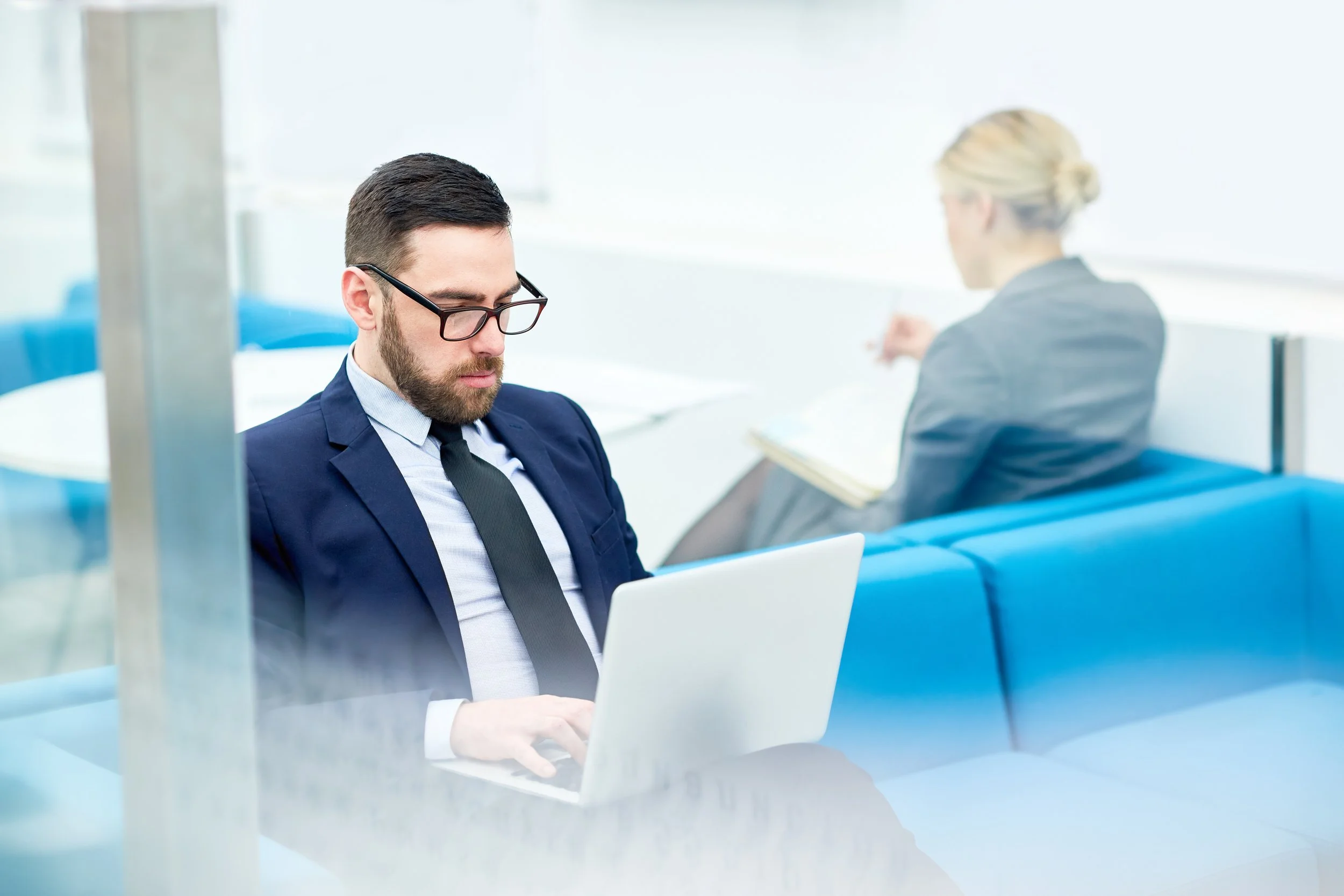 A young man with dark hair, beard, and glasses in a suit working on a laptop in a modern office with another woman in the background.