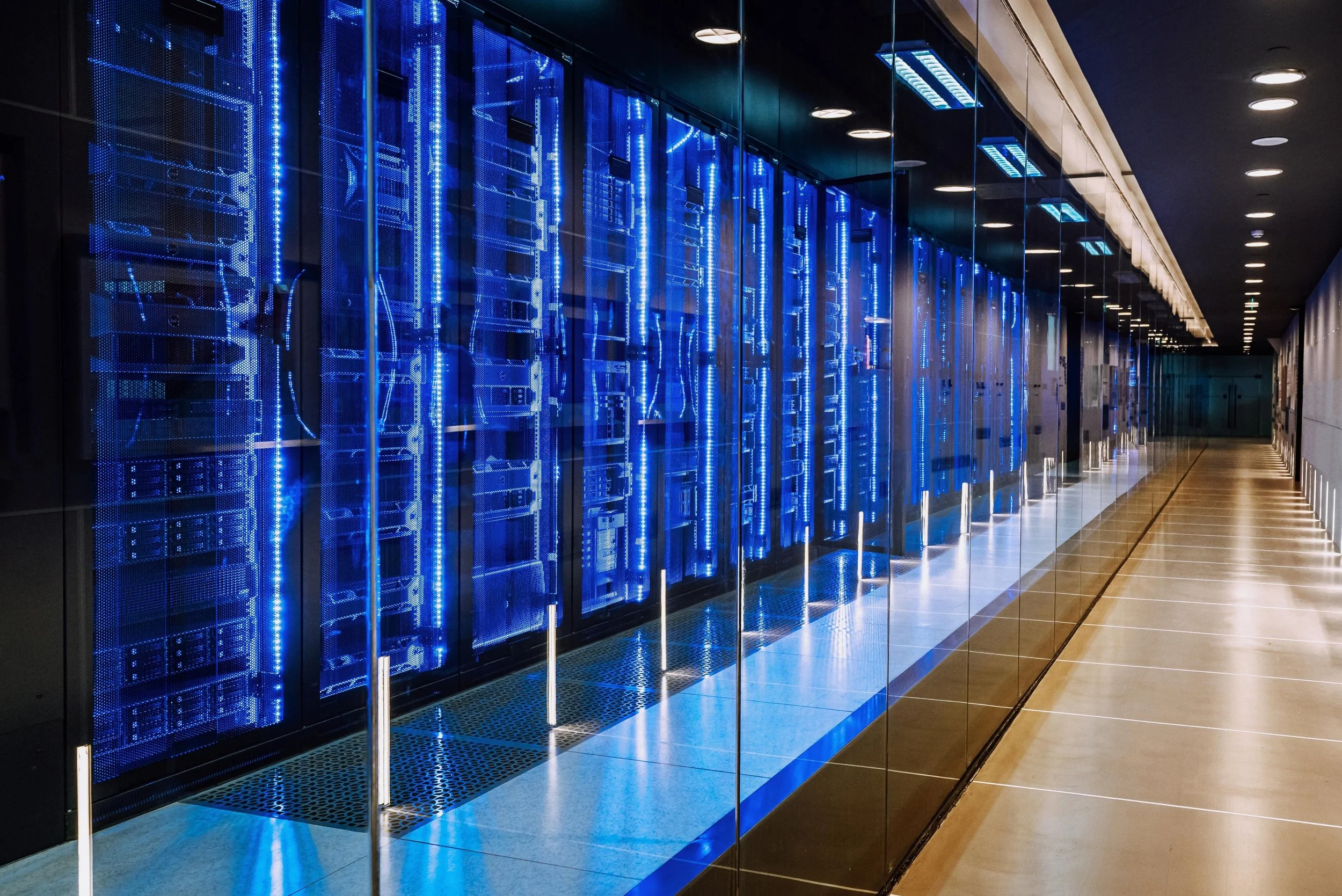 A row of computer servers illuminated by blue LED lights inside a data center, reflected in surrounding glass panels.