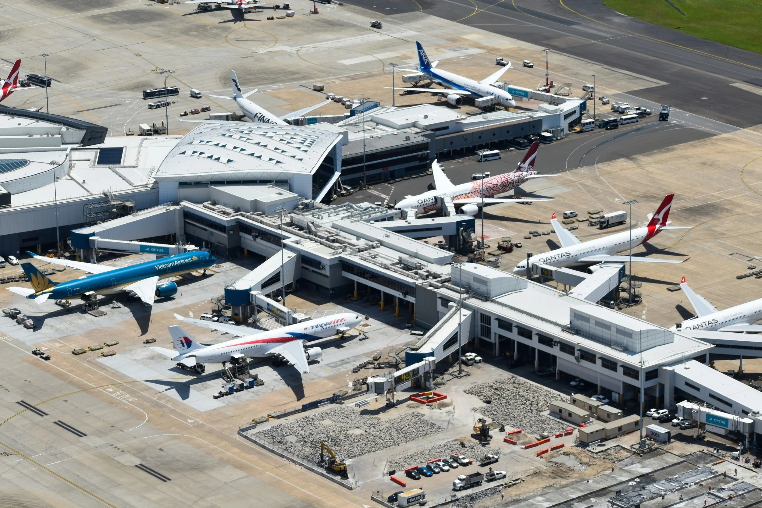 Aerial view of an airport terminal with several airplanes parked at gates, including airlines like Qantas, Malaysia Airlines, Vietnam Airlines, and others. The tarmac shows ground service vehicles, luggage transports, and construction activity with equipment and barriers.
